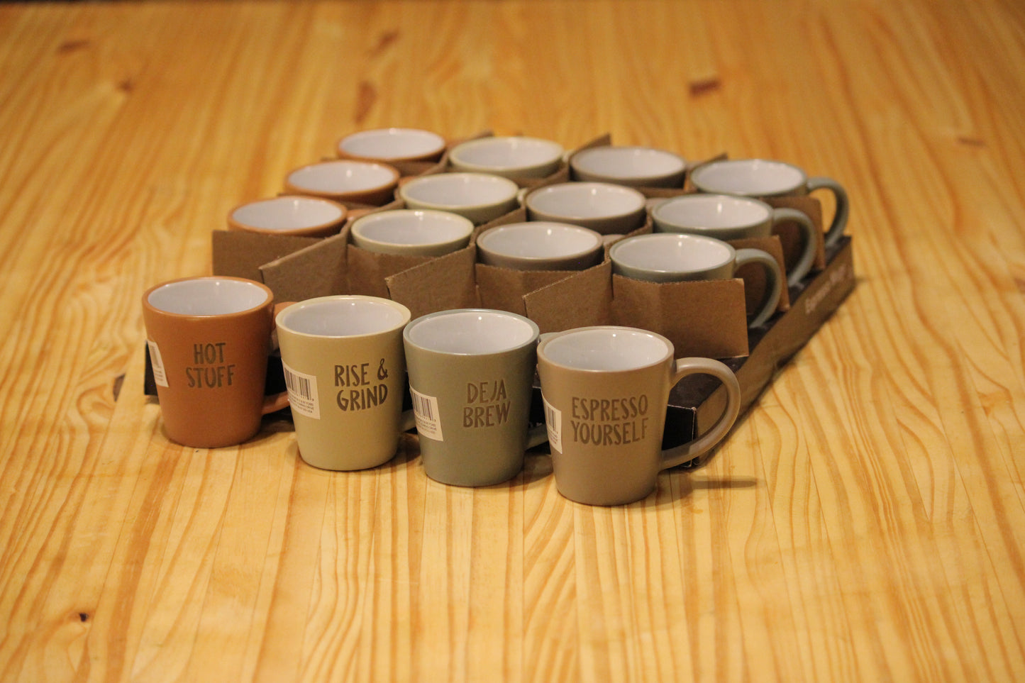 A set of ceramic coffee mugs in various earthy tones, including terracotta, beige, sage green, and taupe, arranged in a cardboard carrier on a light wood tabletop. The front mugs feature printed text labels such as 'HOT STUFF', 'RISE & GRIND', 'DEJA BREW', and 'ESPRESSO YOURSELF'.