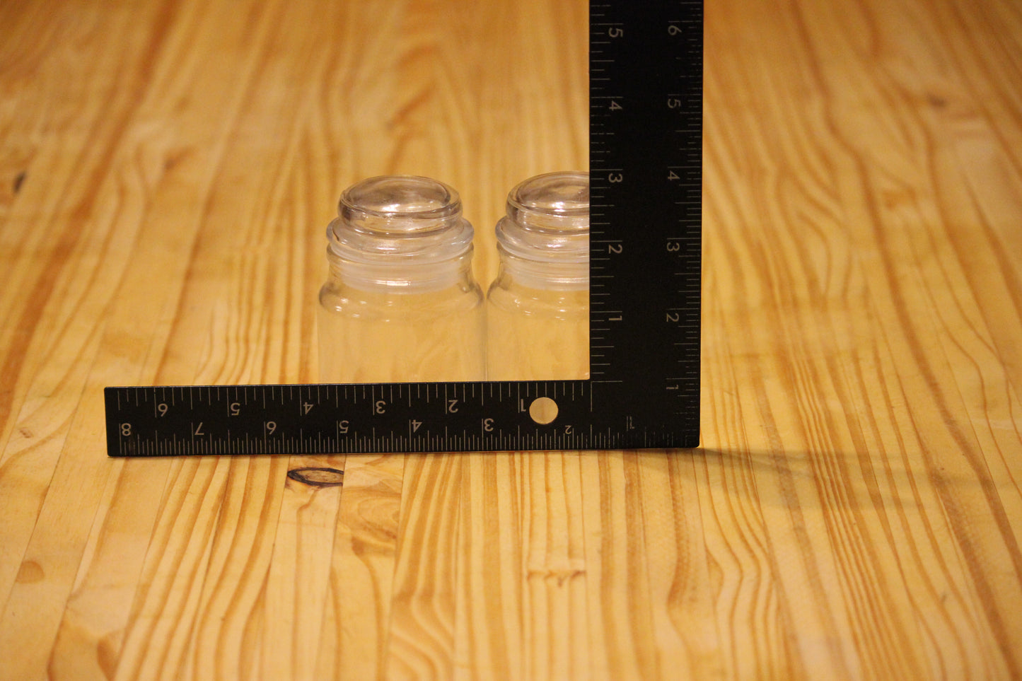 Two small clear glass jars placed on a light wood surface with a black L-square ruler showing measurements in centimeters and inches.