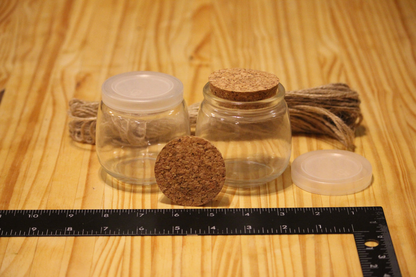 Two small clear glass jars with lids on a wooden surface, one with a cork lid and one with a translucent lid, alongside a cork stopper and a black ruler on a wood-grain background.