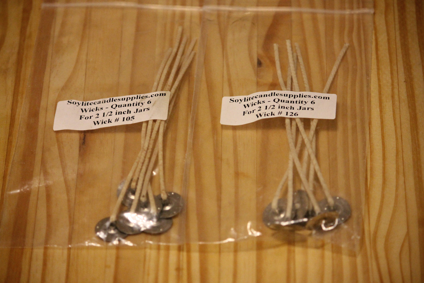 Two clear plastic bags containing Wick #105 and Wick #126 soy candle wicks with silver metal wick stands, labeled with a white paper tag showing the brand 'Soyflitecandlesupplies.com', placed on a light wood background.