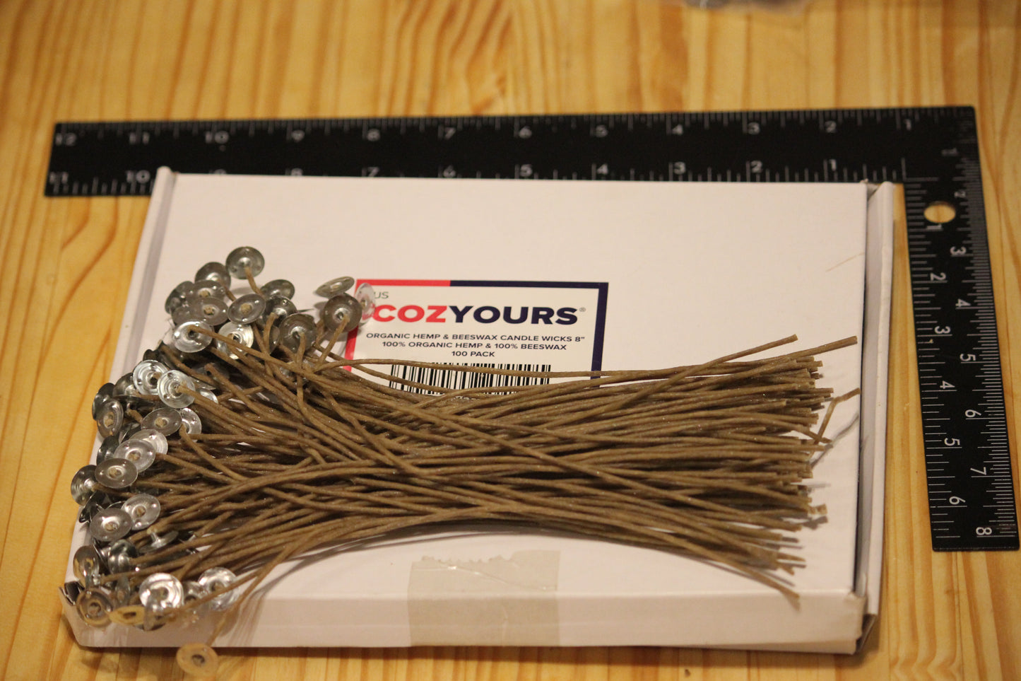 A box of CozYours candle wicks with metal wick caps, displayed on a wooden table next to a black ruler for scale.