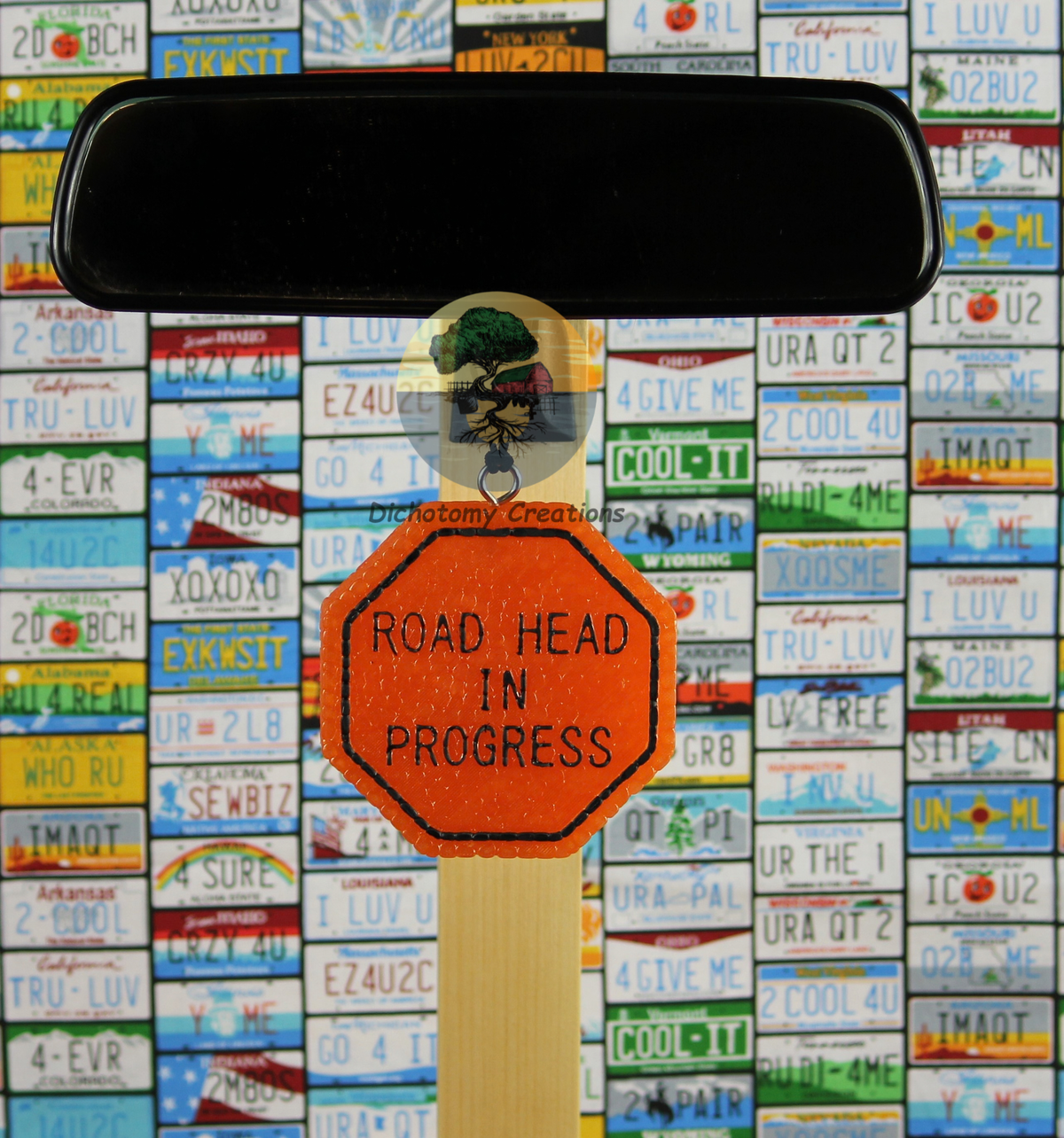 A orange road sign freshie with the words 'ROAD HEAD IN PROGRESS', displayed on a rearview mirror in front of a colorful background of license plates.