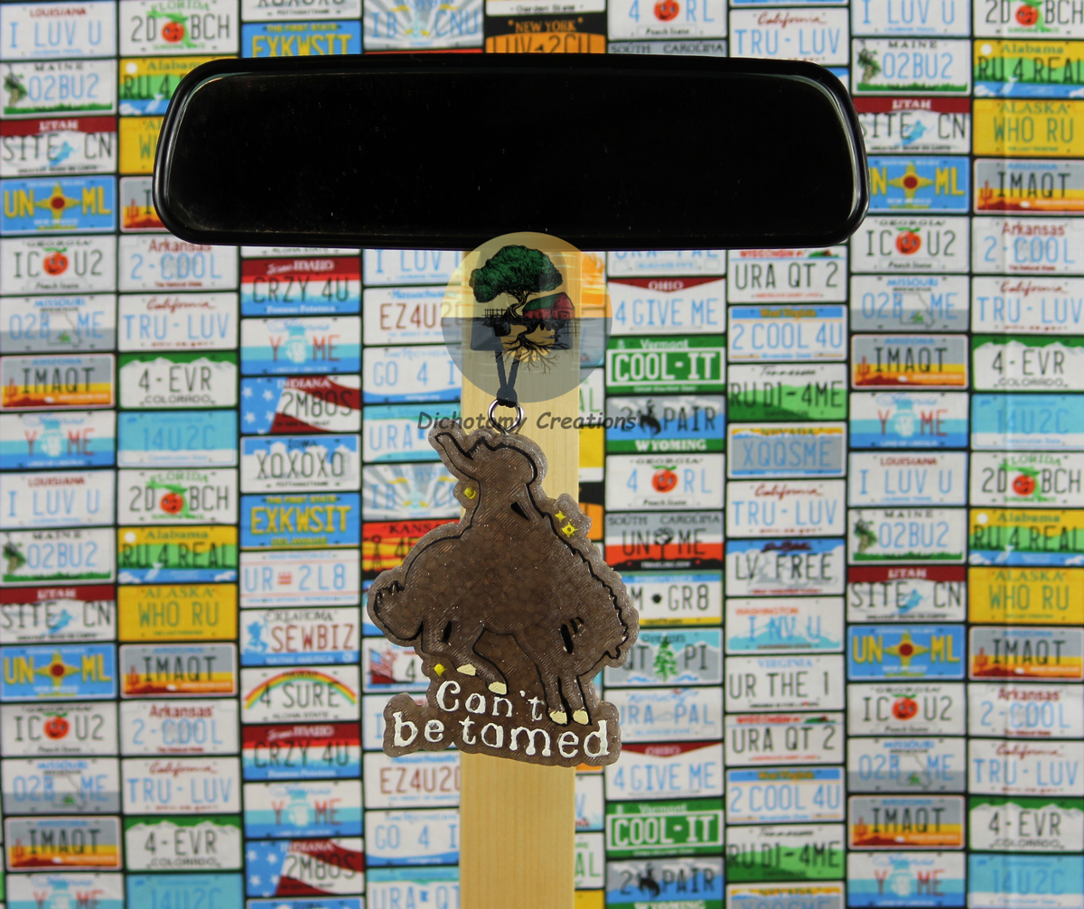 A brown freshie showing a silhouette of a woman riding a bucking bronco that reads 'CAN'T BE TAMED', displayed on a rearview mirror in front of a colorful background of license plates.