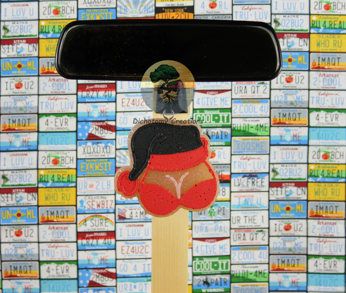 A beige freshie, shaped as boobs in a red bra with a red and black santa hat. Displayed from a rearview mirror with a background of colorful license plates.