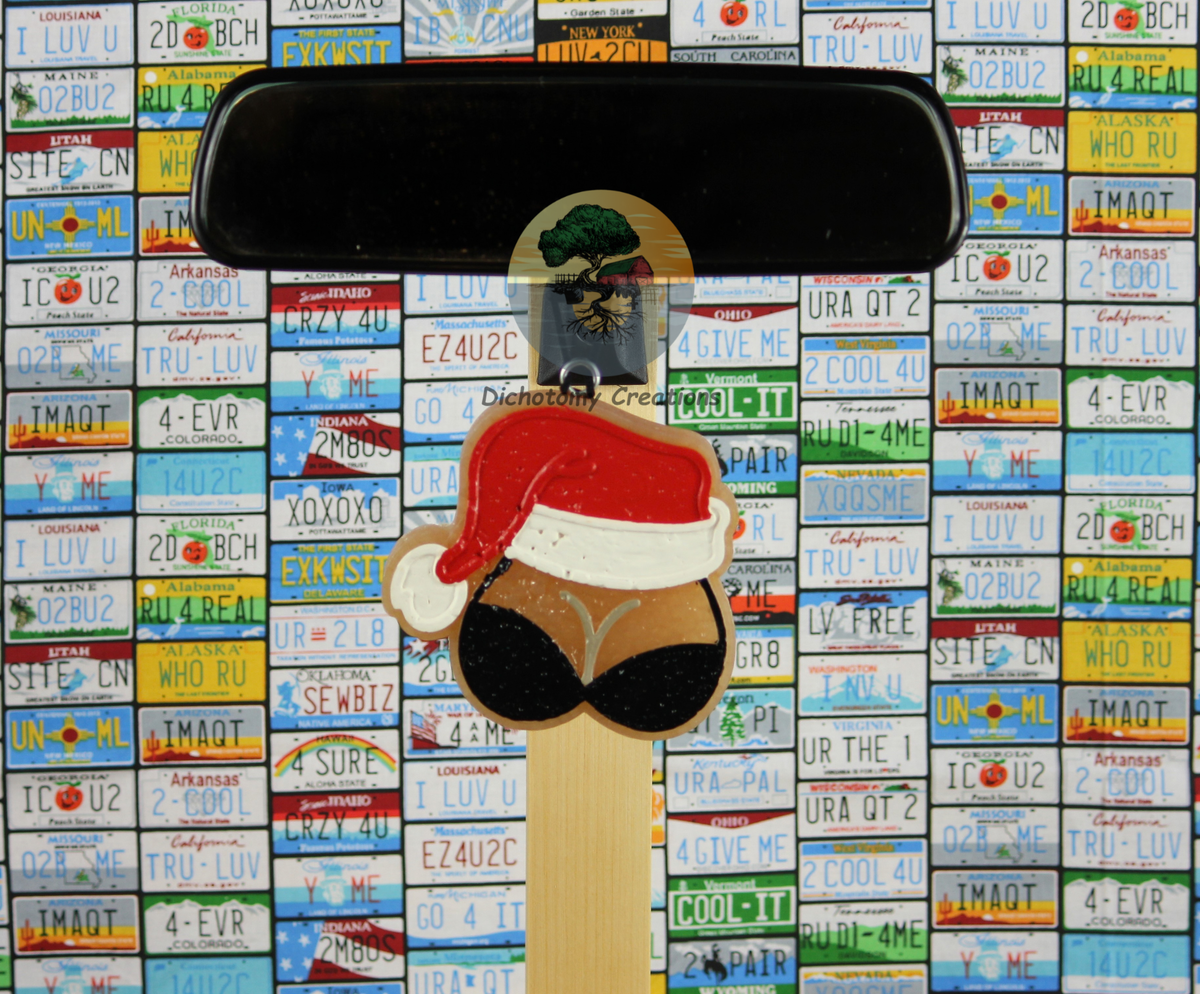A beige freshie, shaped as boobs in a black bra with a red and white santa hat. Displayed from a rearview mirror with a background of colorful license plates.