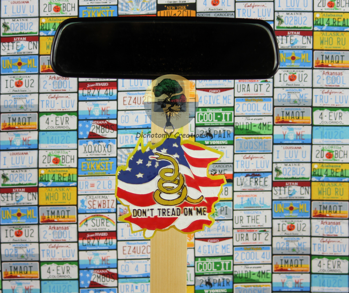 A yellow freshie with a snake and the words 'DON'T TREAD ON ME' with an American flag, displayed on a rearview mirror, set against a background of colorful license plates.