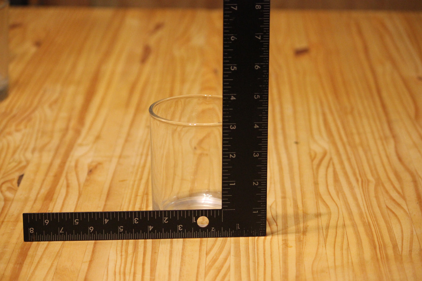 A clear glass tumbler on a light wood tabletop, with a black L-square ruler positioned behind it to show its height and width measurements.