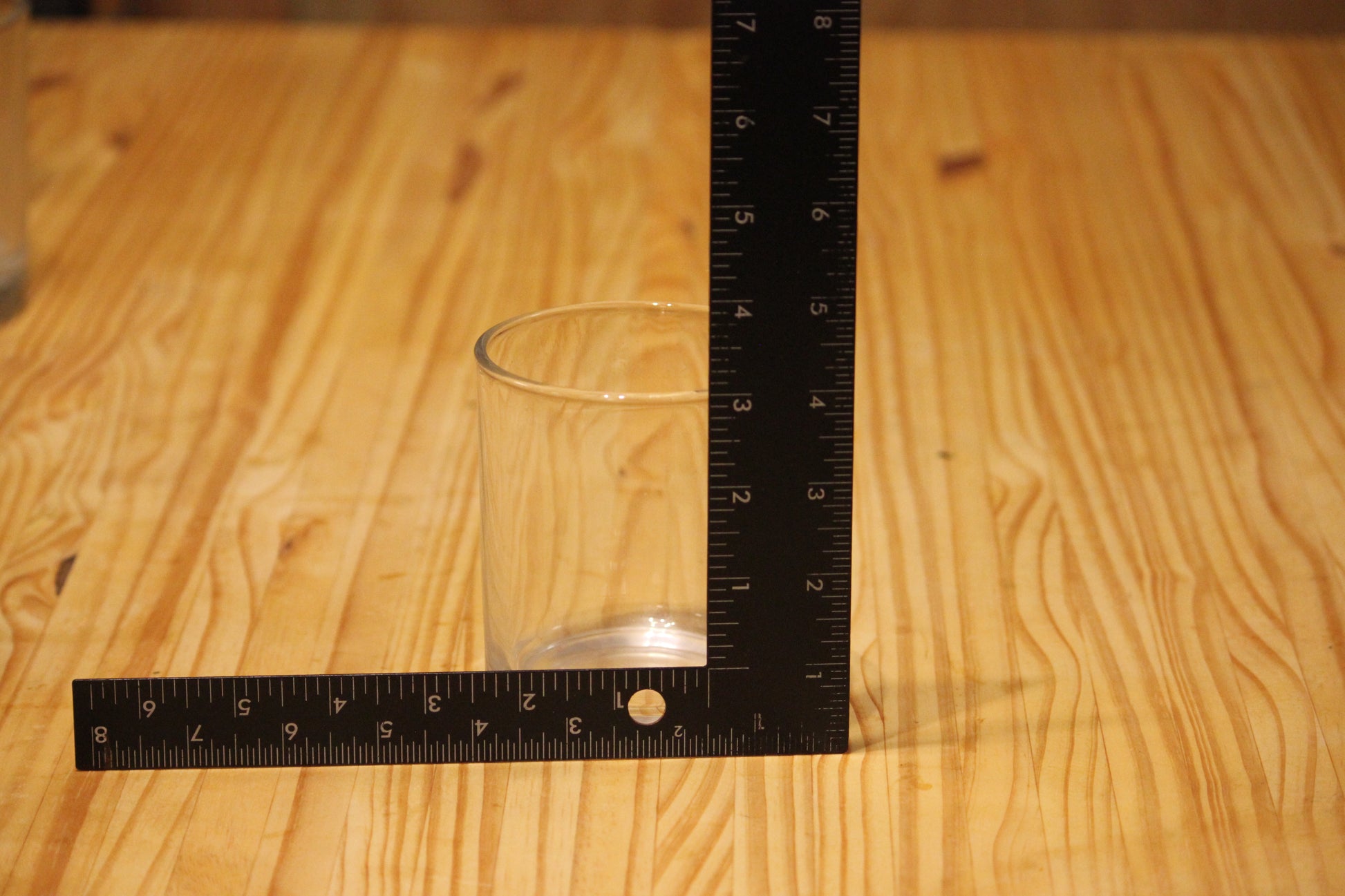 A clear glass tumbler on a light wood tabletop, with a black L-square ruler positioned behind it to show its height and width measurements.