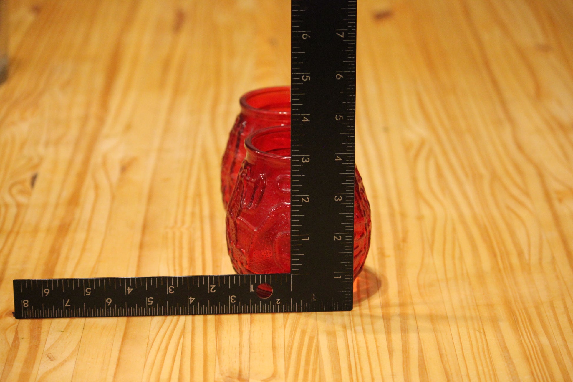 A red glass jar candle holder with a textured pattern, shown next to a black L-square ruler on a light wood grain surface to indicate its size.
