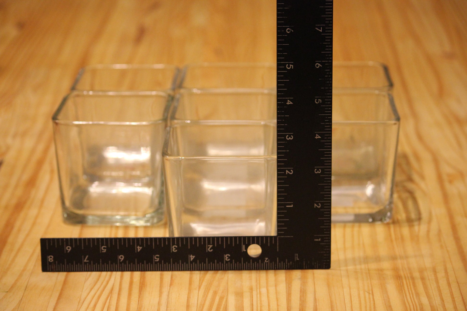 Seven clear square glass cups on a wooden surface, with a black ruler to show their size.