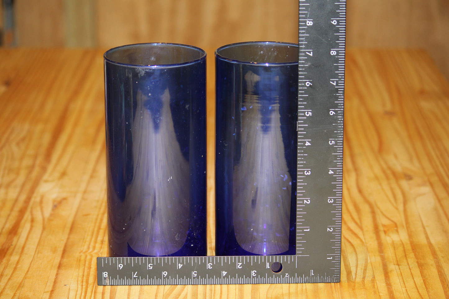 Two translucent dark blue cylindrical vessels standing on a wooden table, with metal rulers placed at the bottom and along the right side for scale.