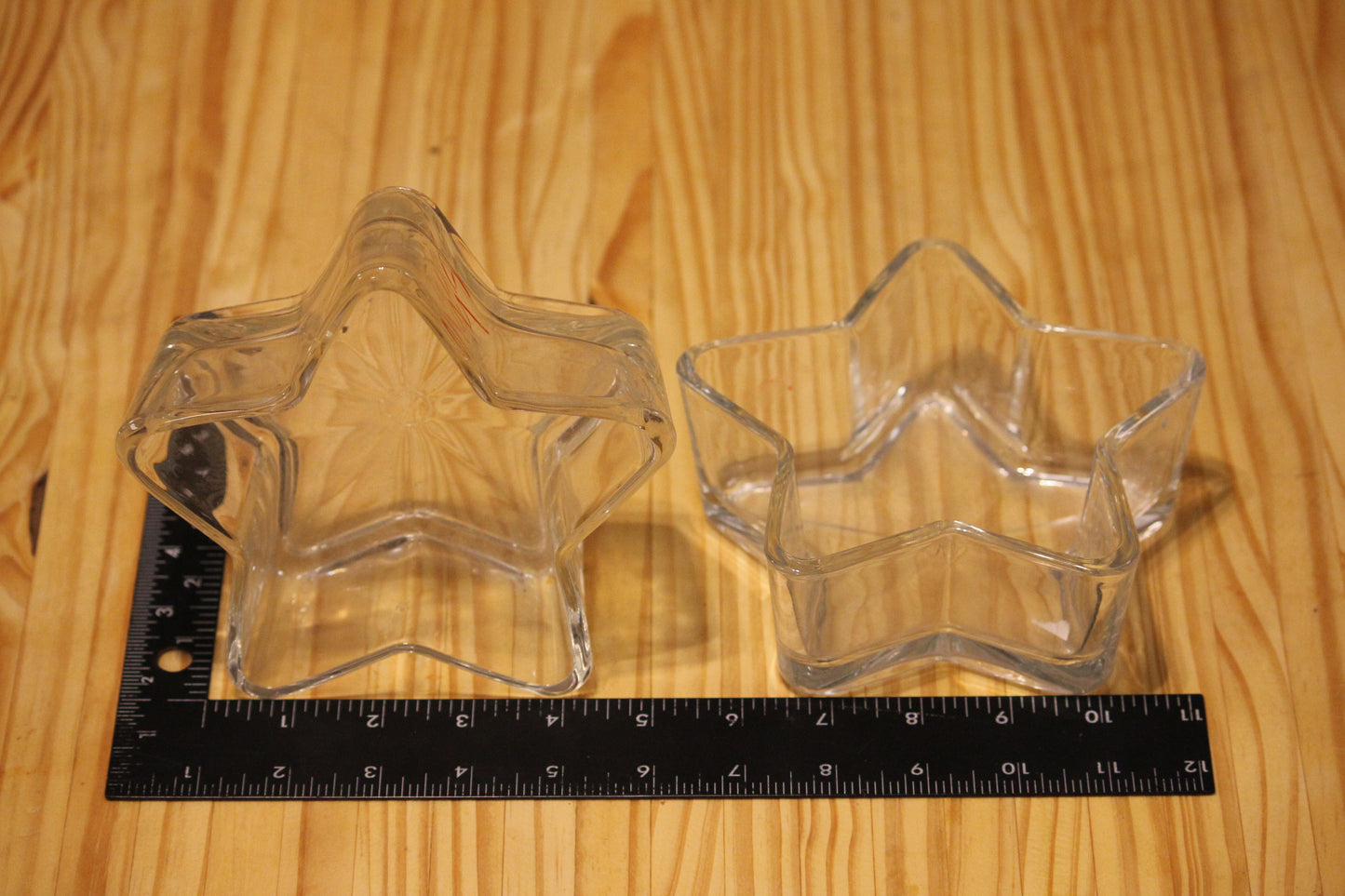 Two clear star-shaped glass vessels shown on a light wood surface, with a black ruler placed underneath for size reference.