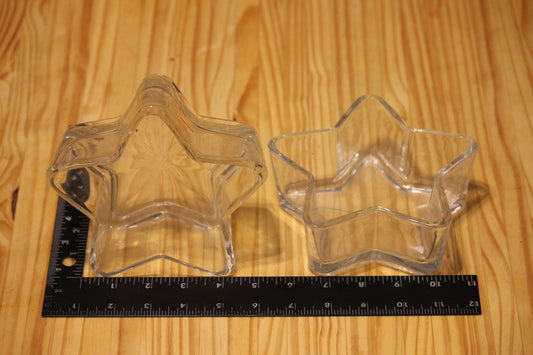 Two clear star-shaped glass vessels shown on a light wood surface, with a black ruler placed underneath for size reference.