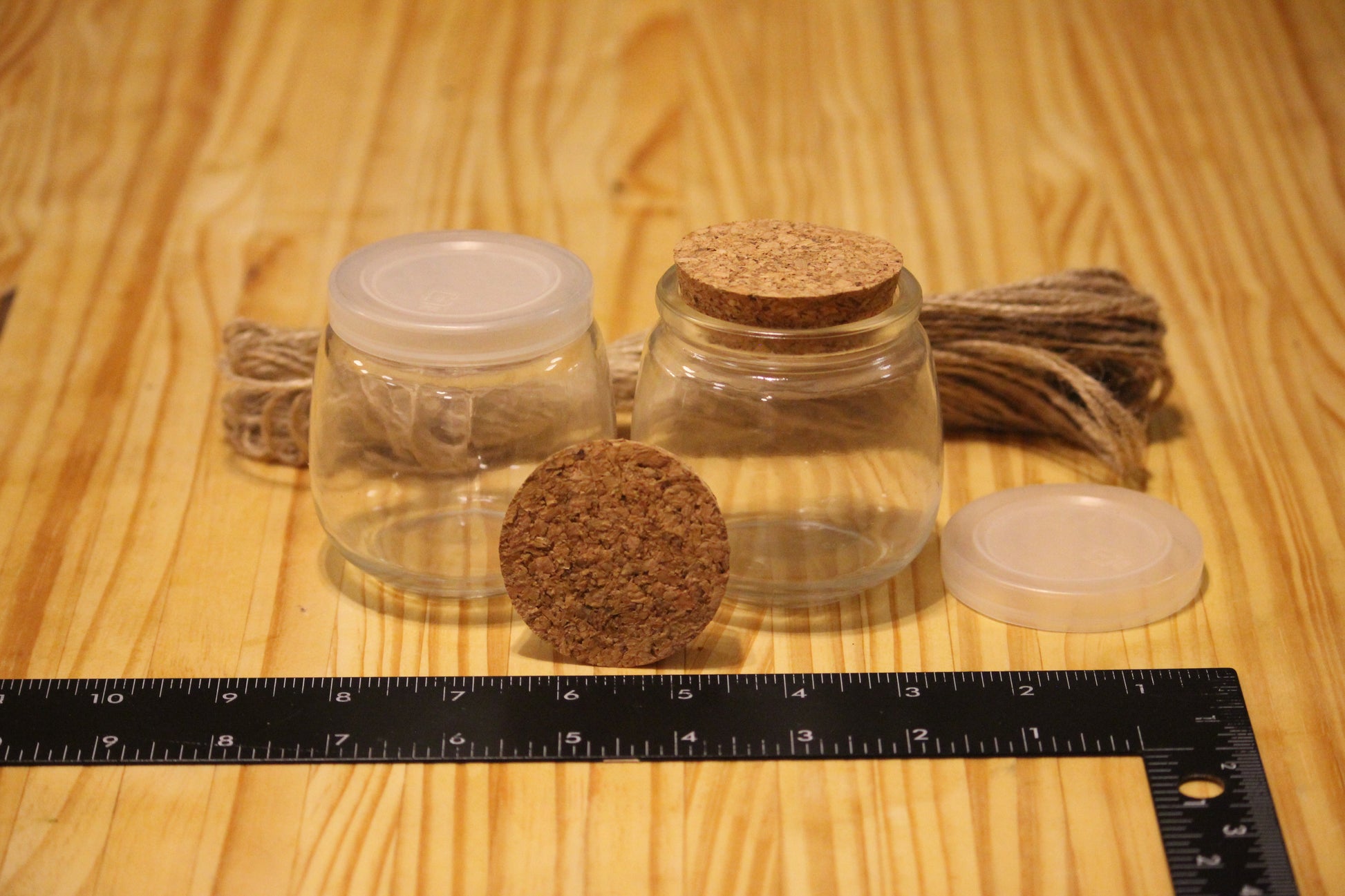 Two small clear glass jars with lids on a wooden surface, one with a cork lid and one with a translucent lid, alongside a cork stopper and a black ruler on a wood-grain background.