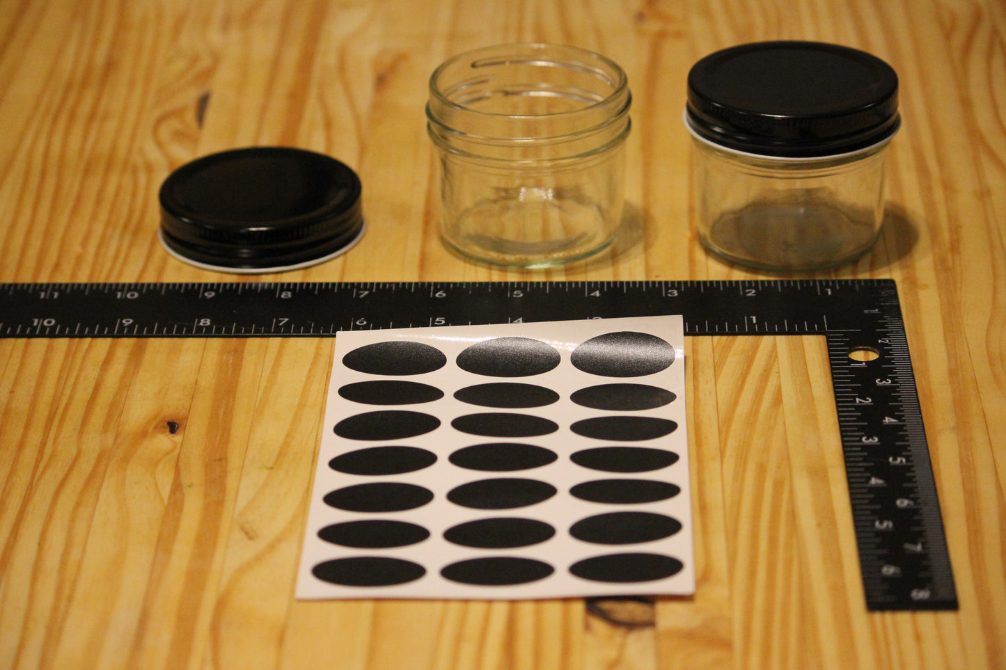 Two small clear glass jars with black lids and a sheet of oval black labels on a wooden table, with a ruler and L-square measuring the labels and jars.