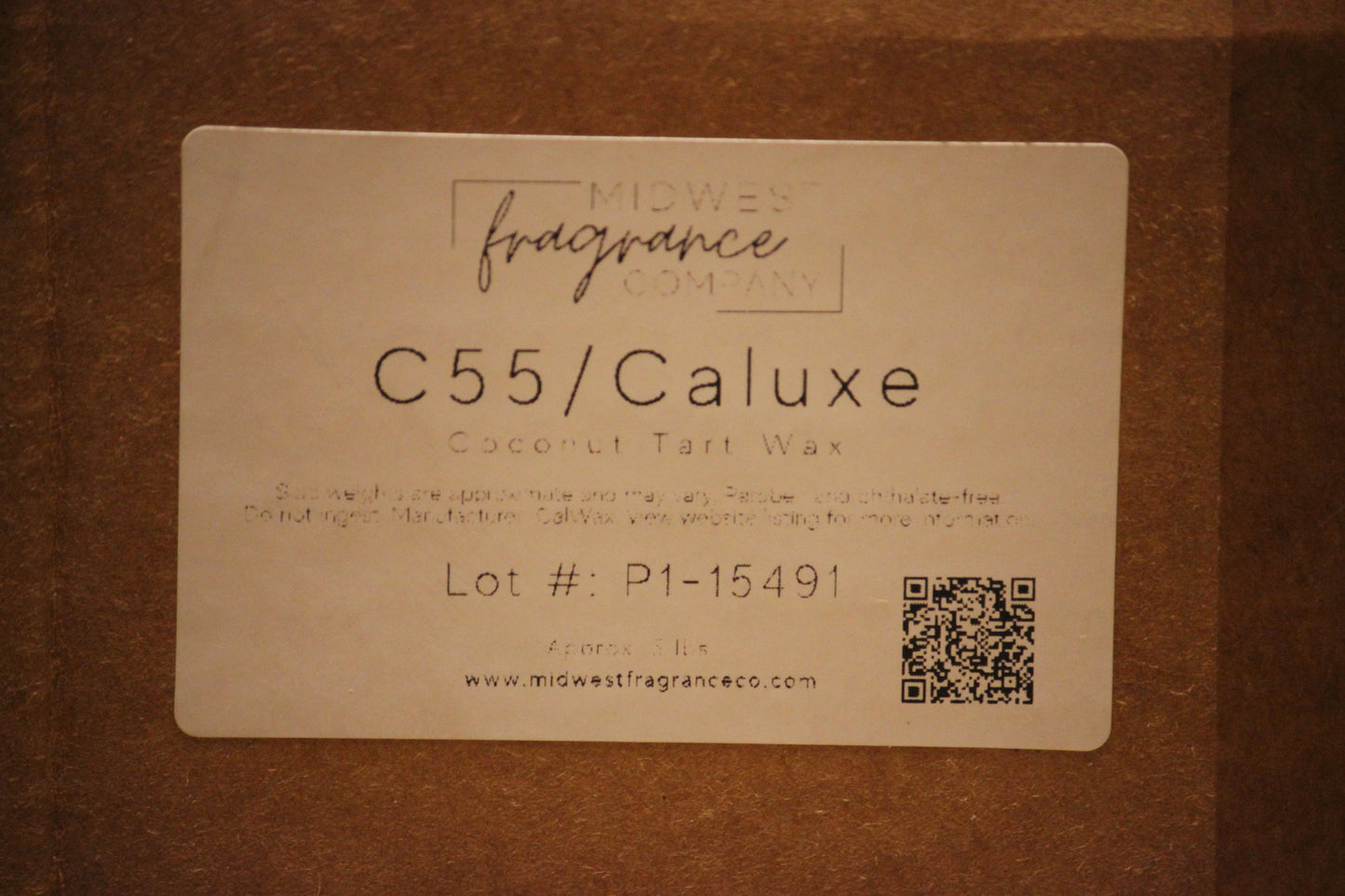 A square paper label for MidWest Fragrance Company's C55/Caluxxe Coconut Tart Wax, displayed on a brown cardboard background.