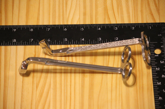 Candle wick trimmers, silver, placed on a light wood surface next to a black measuring ruler for scale.