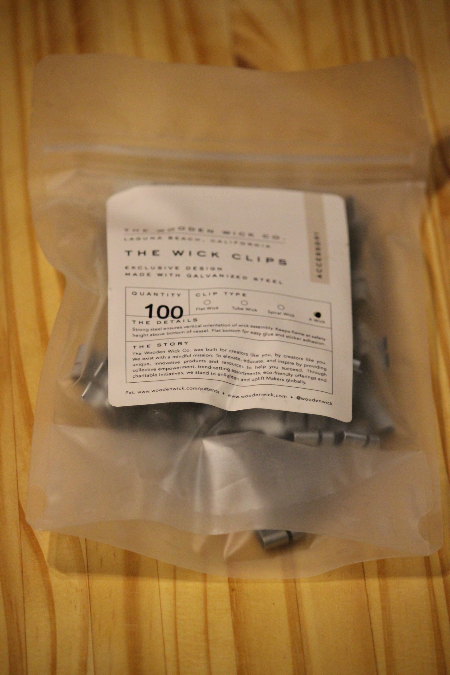 A clear plastic bag containing X wick clips with a white paper label reading 'THE WICK CLIPS' and '100', placed on a light wood surface.