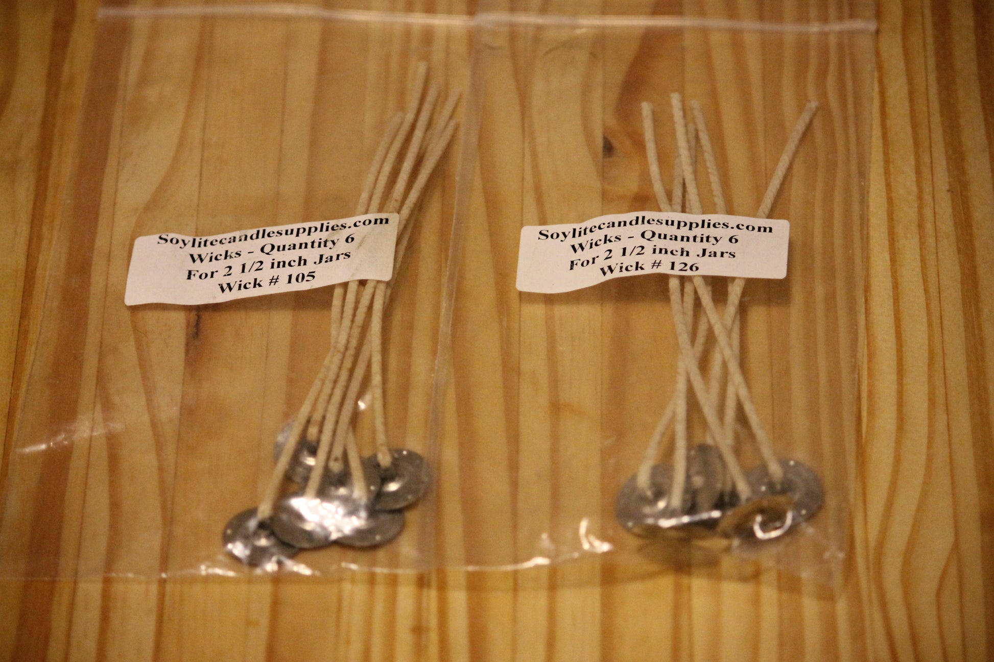 Two clear plastic bags containing Wick #105 and Wick #126 soy candle wicks with silver metal wick stands, labeled with a white paper tag showing the brand 'Soyflitecandlesupplies.com', placed on a light wood background.