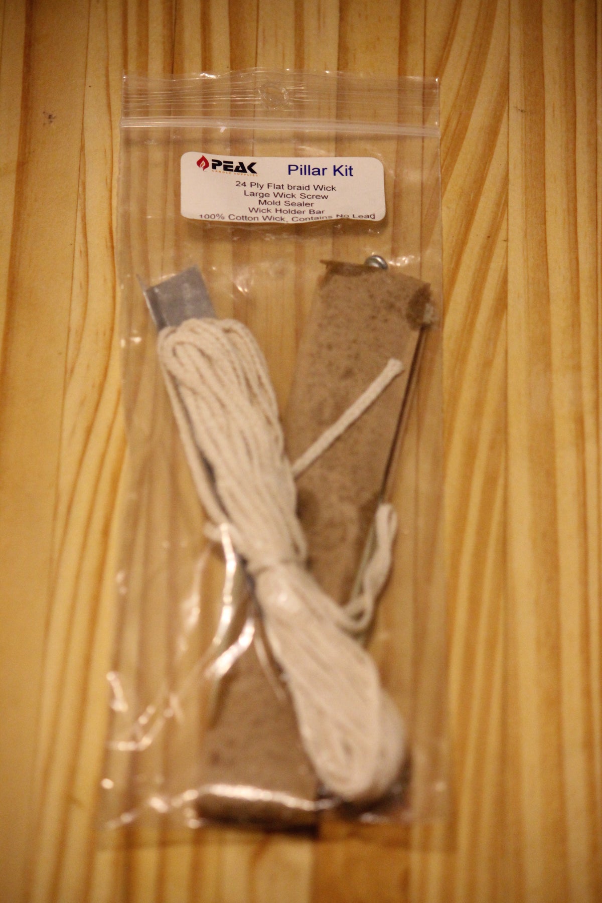 A packaged 'Pillar Kit' by Peak, containing a bundle of cotton wick and accessories, sealed in a clear plastic bag and placed on a wooden surface.