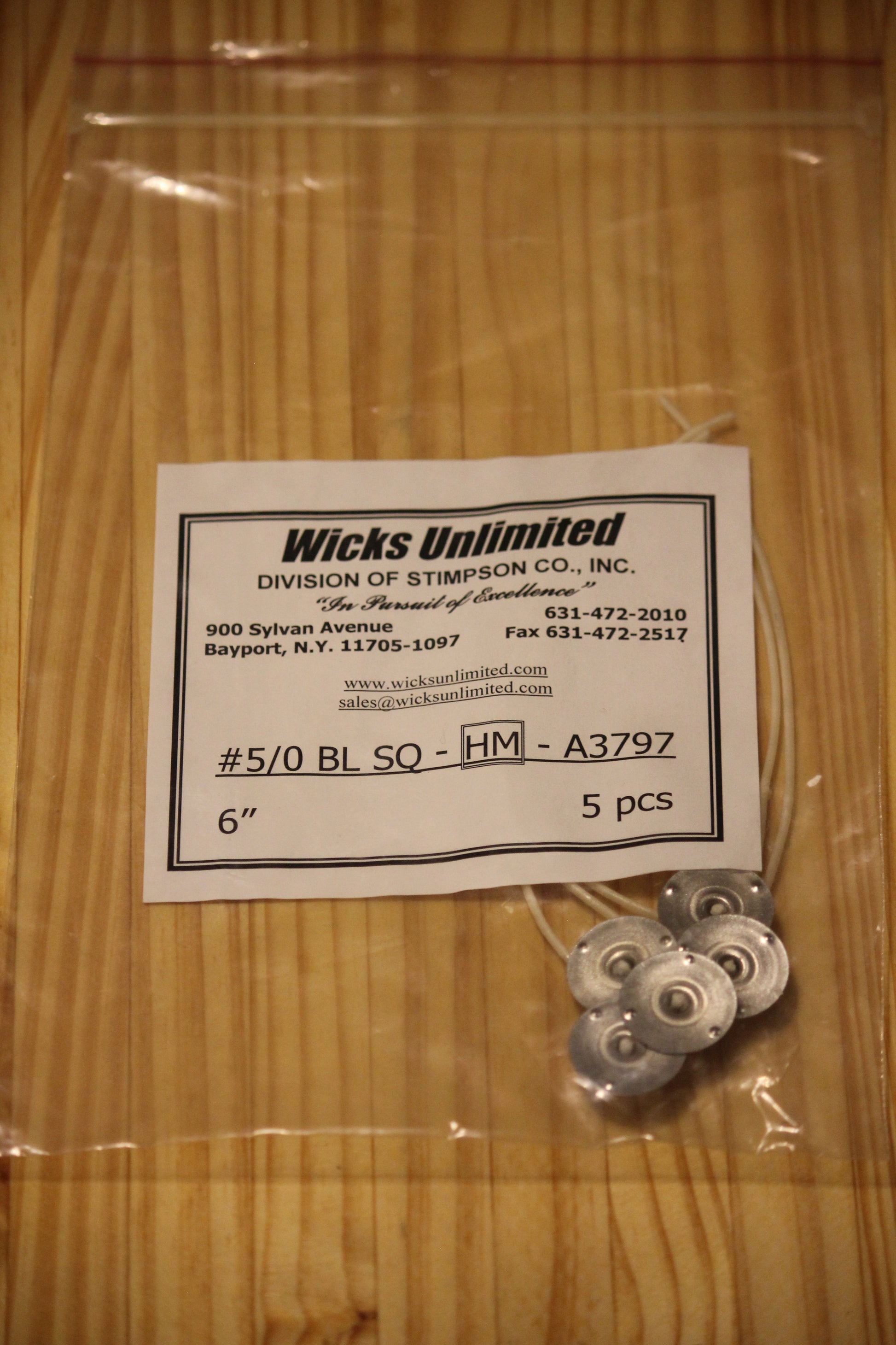 A clear plastic zip-top bag containing five six inch pre-tabbed wicks next to a white paper label that reads 'Wicks Unlimited' and shows product details, placed on a light wood surface.