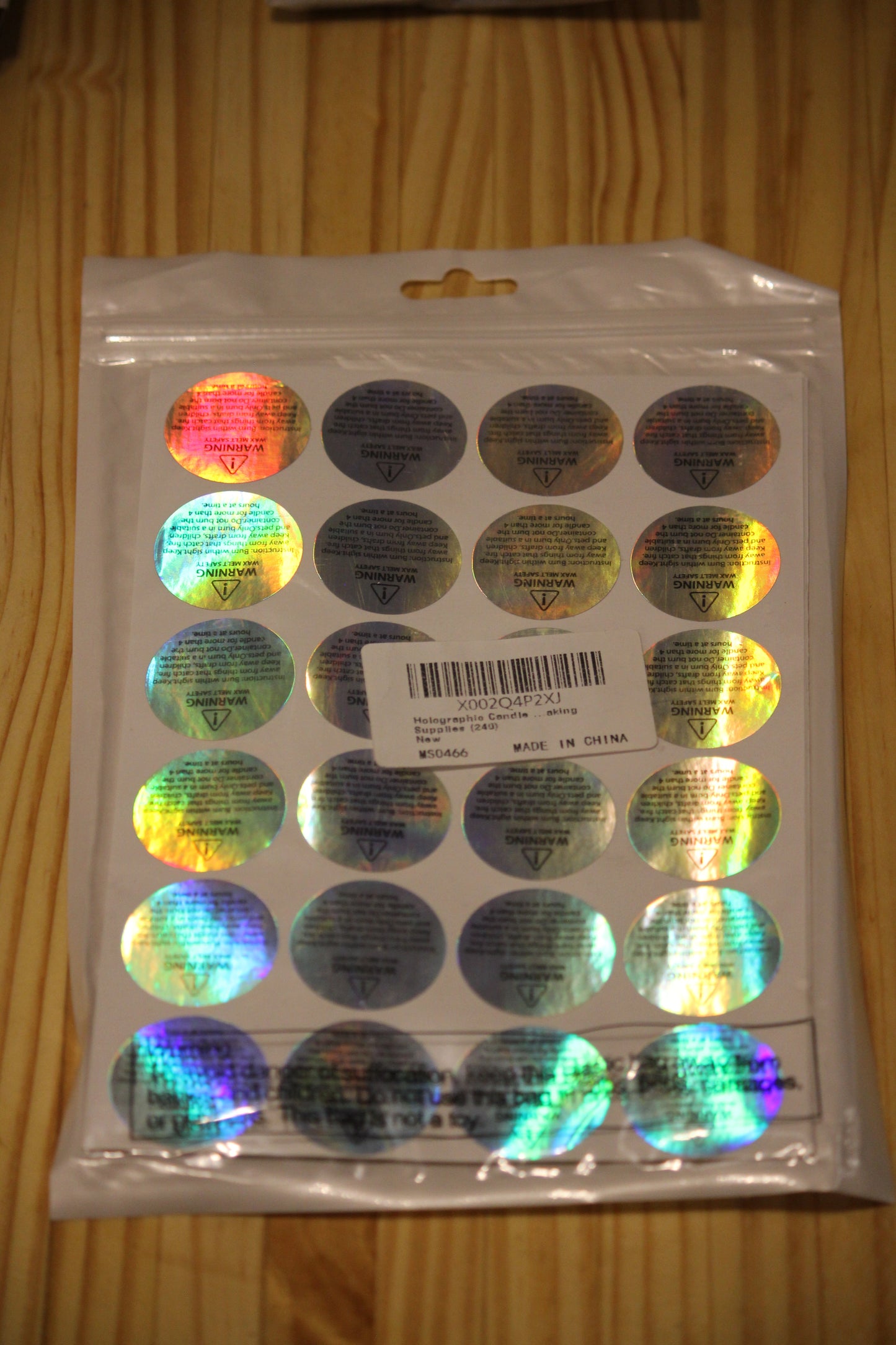 A clear plastic bag containing a sheet of round holographic wax melt warning stickers on a wooden surface. 