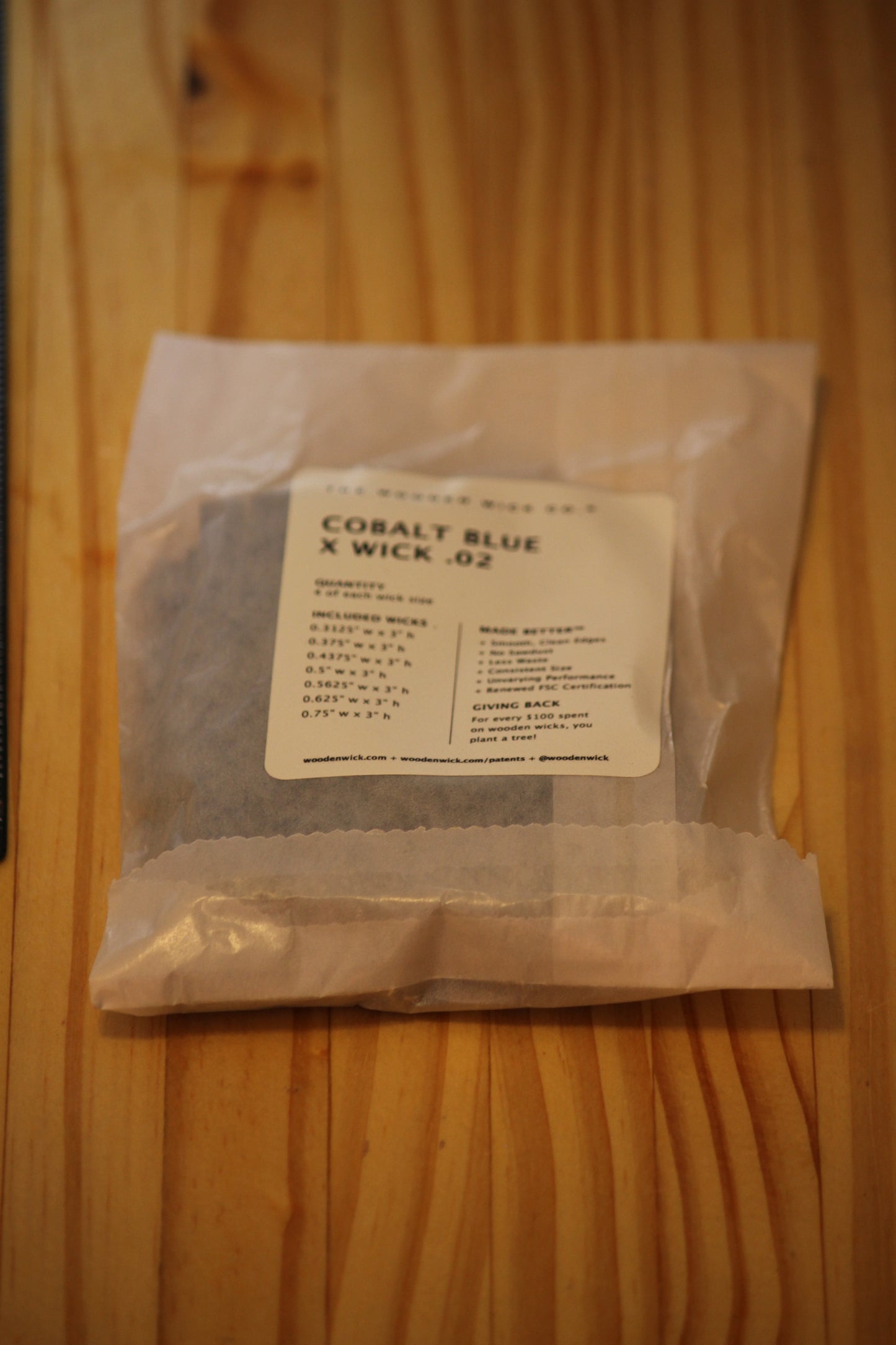 A clear plastic bag containing dark blue wood wick material with a white label reading 'COBALT BLUE X WICK .02', placed on a light wood grain surface.