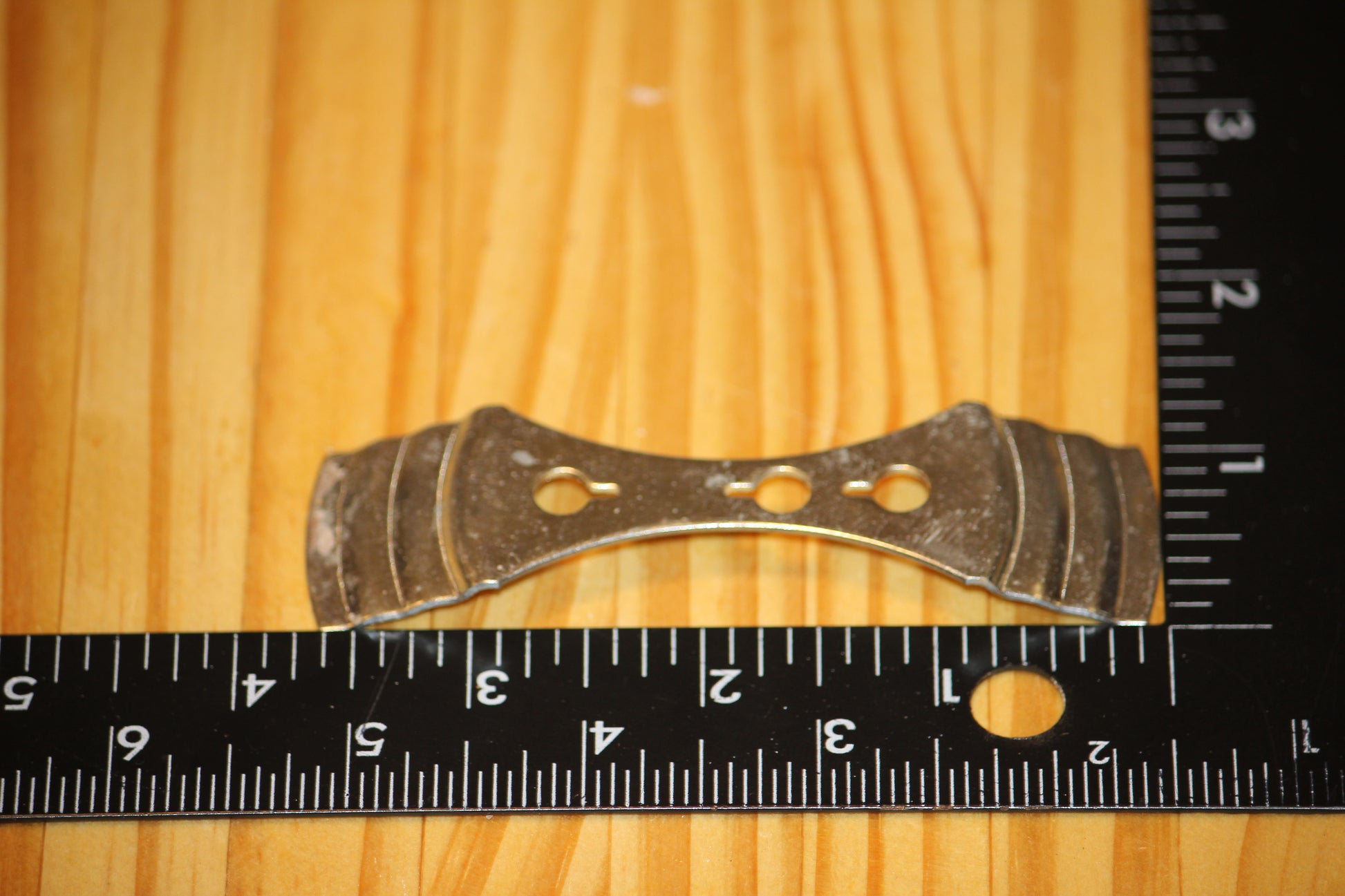 A small metal wick centering device, shown on a light wood grain surface with black rulers for scale.