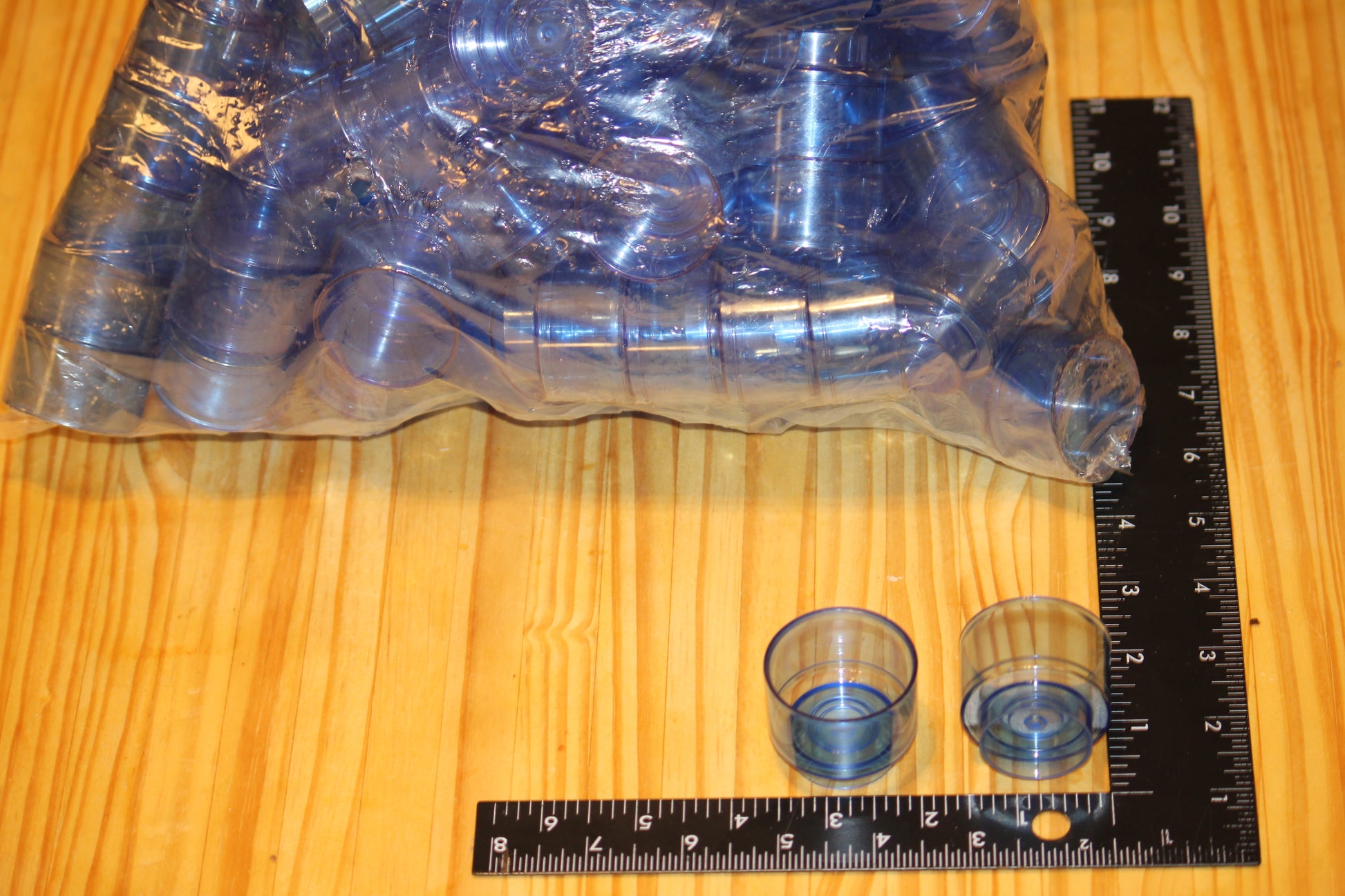 A bulk pack of clear plastic tea light  cups, wrapped in protective plastic, displayed on a wooden surface next to black rulers for scale.