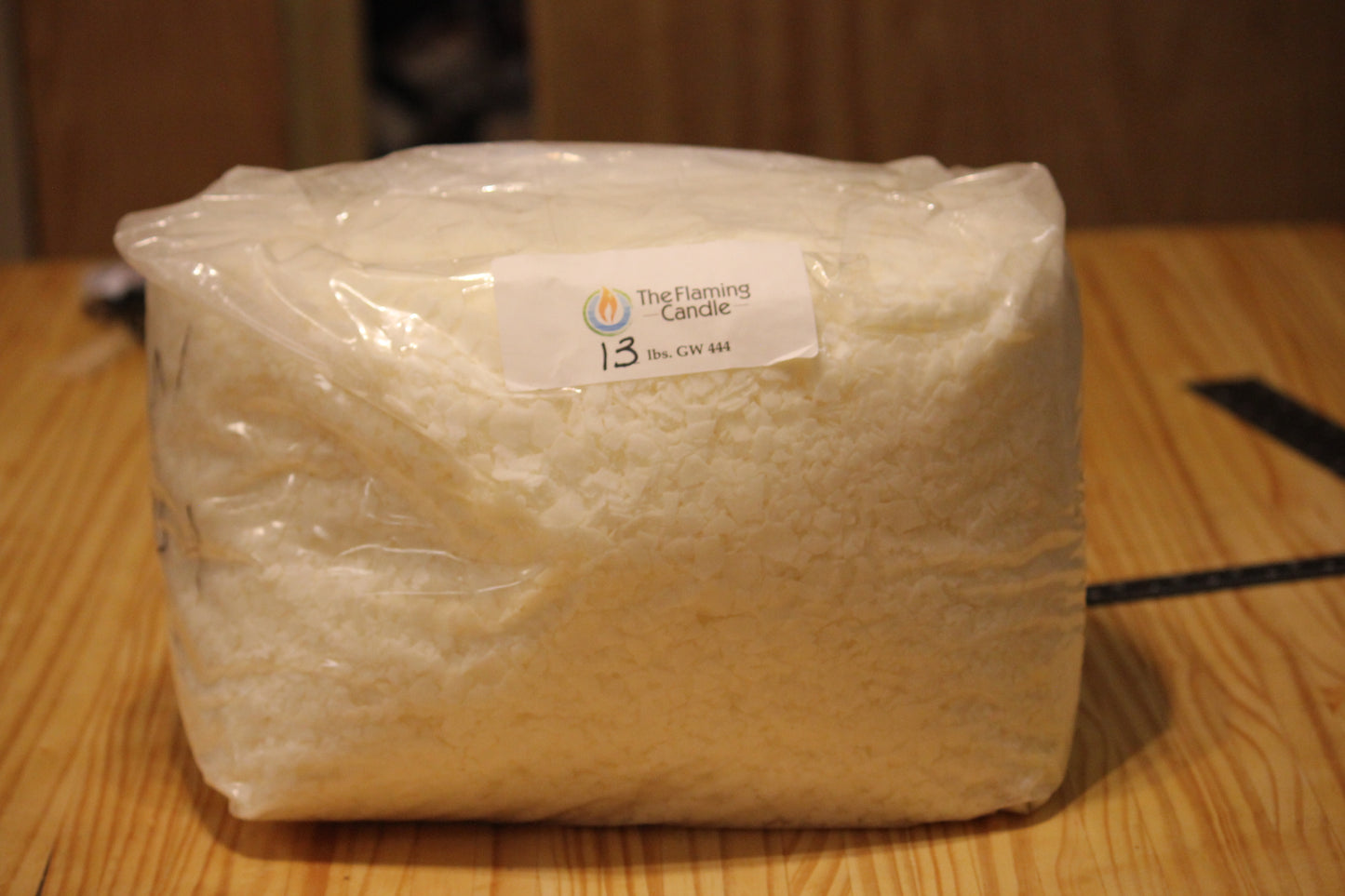 A clear plastic bag filled with wax flakes, sealed with a white paper label that reads 'The Flaming Candle GW 444', sitting on a wooden tabletop.