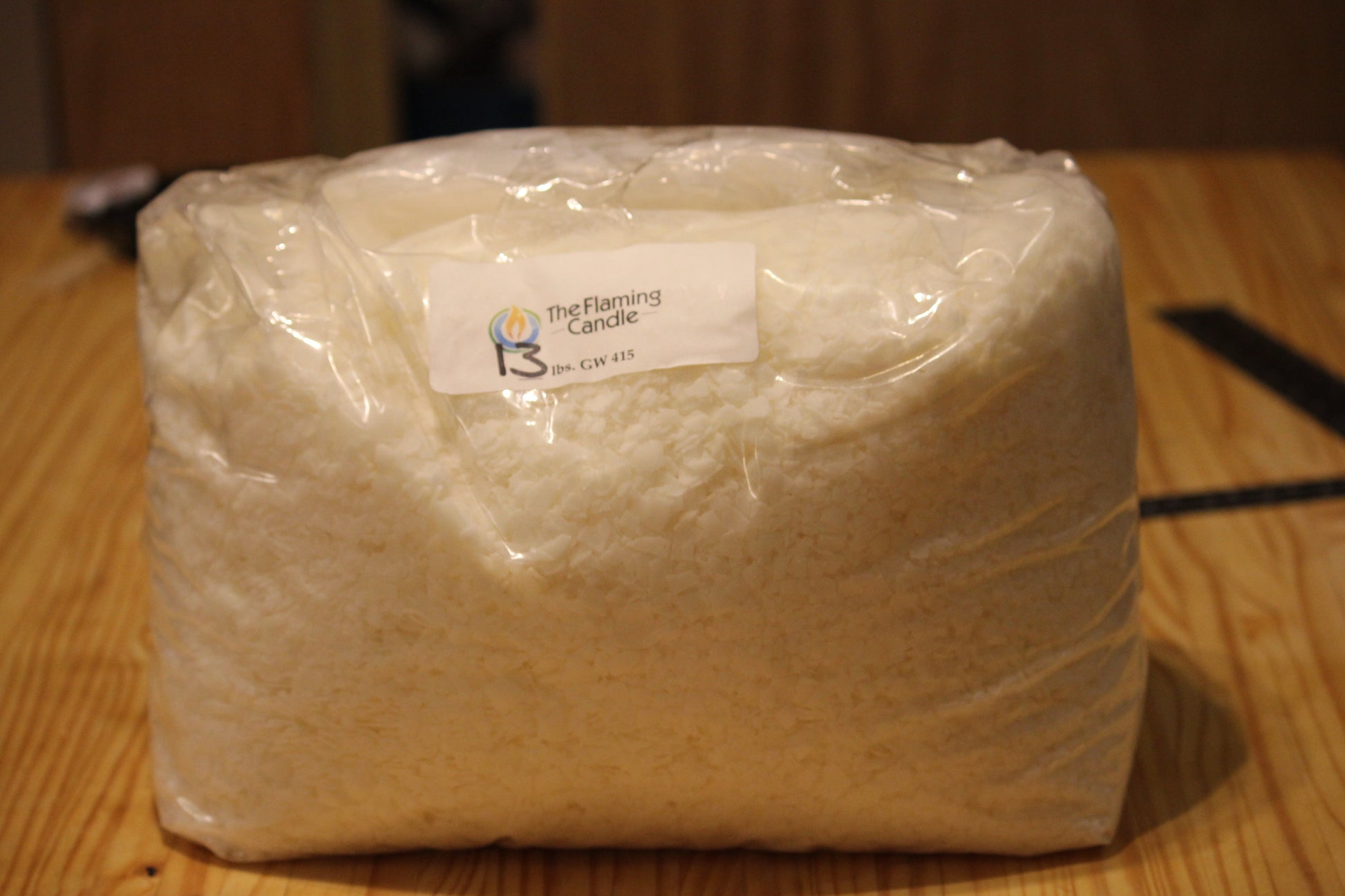 A large bulk package of white wax candle material wrapped in clear plastic, with a white label reading 'The Flaming Candle' and 'GW 415', sitting on a wooden tabletop.