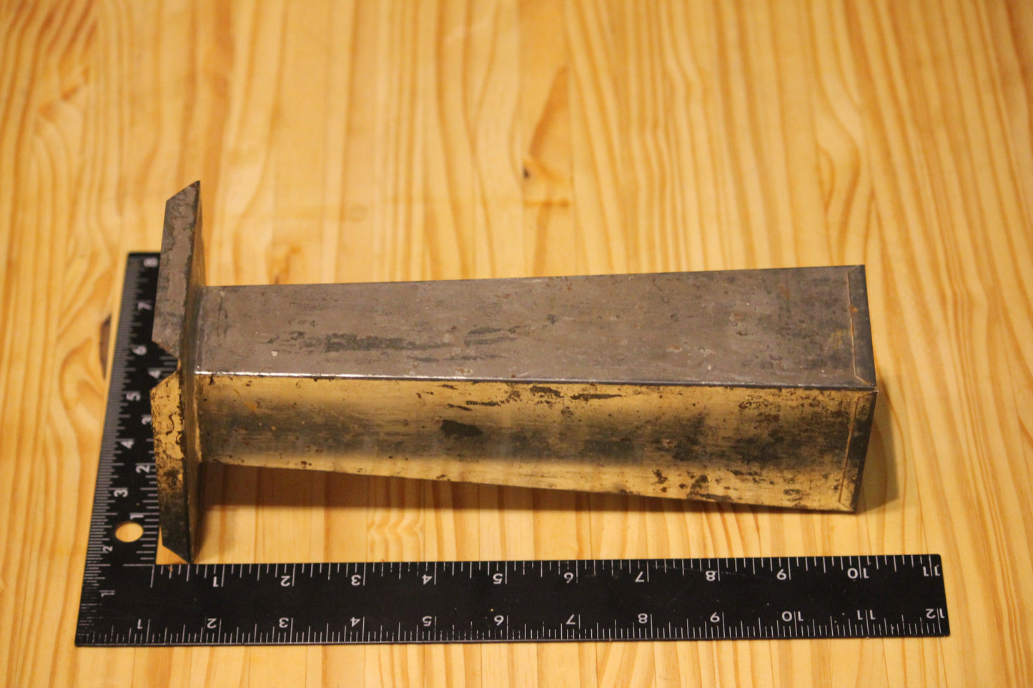 A metal pillar candle mold, shown on a light wood grain surface next to a black ruler for scale.
