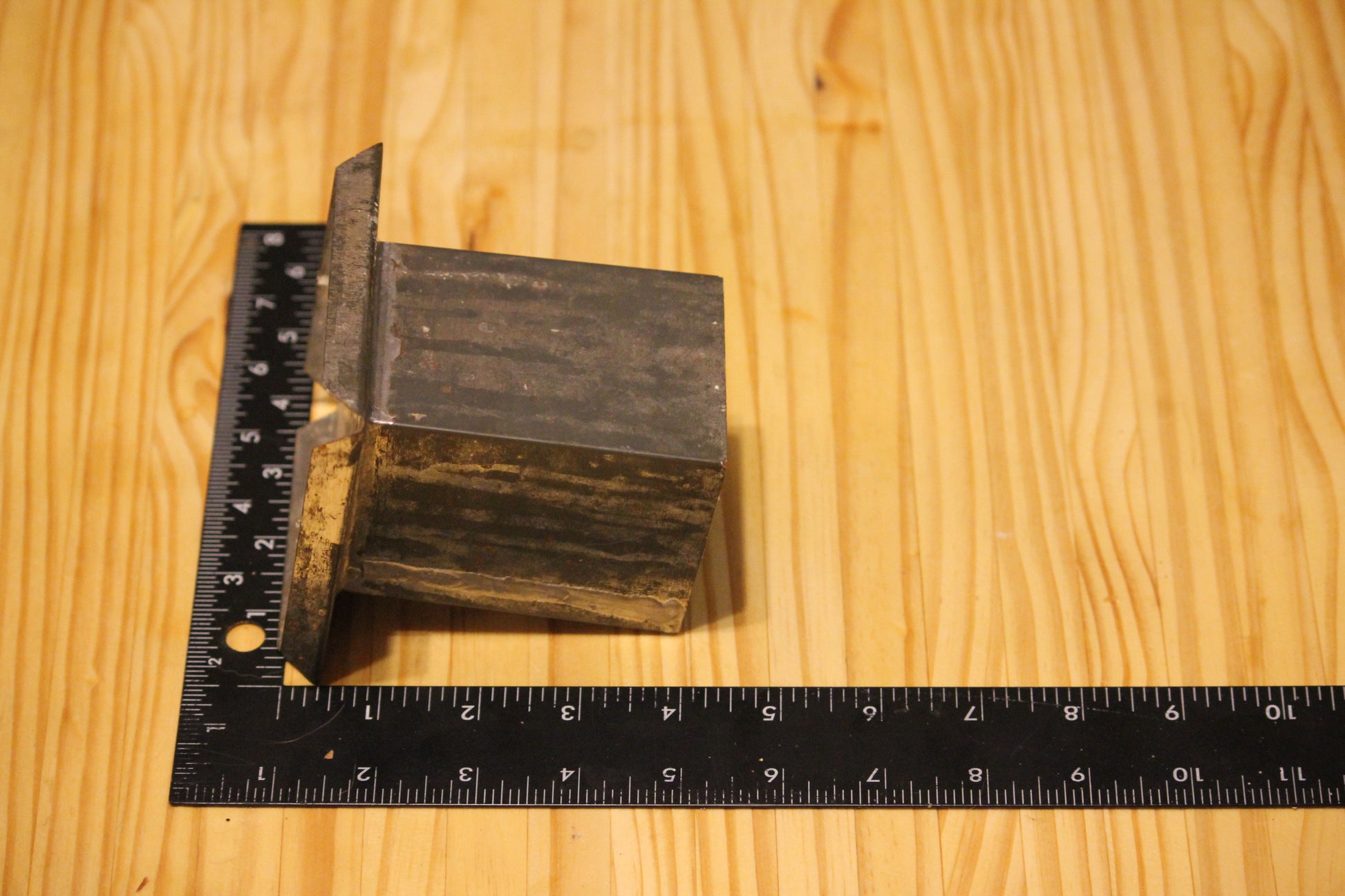A metal square pillar mold, shown on a light wood grain background with a black ruler for scale.