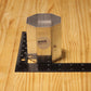 A small metallic octagonal block with a circular label reading 'M2026' stands on a black ruler on a light wood tabletop.