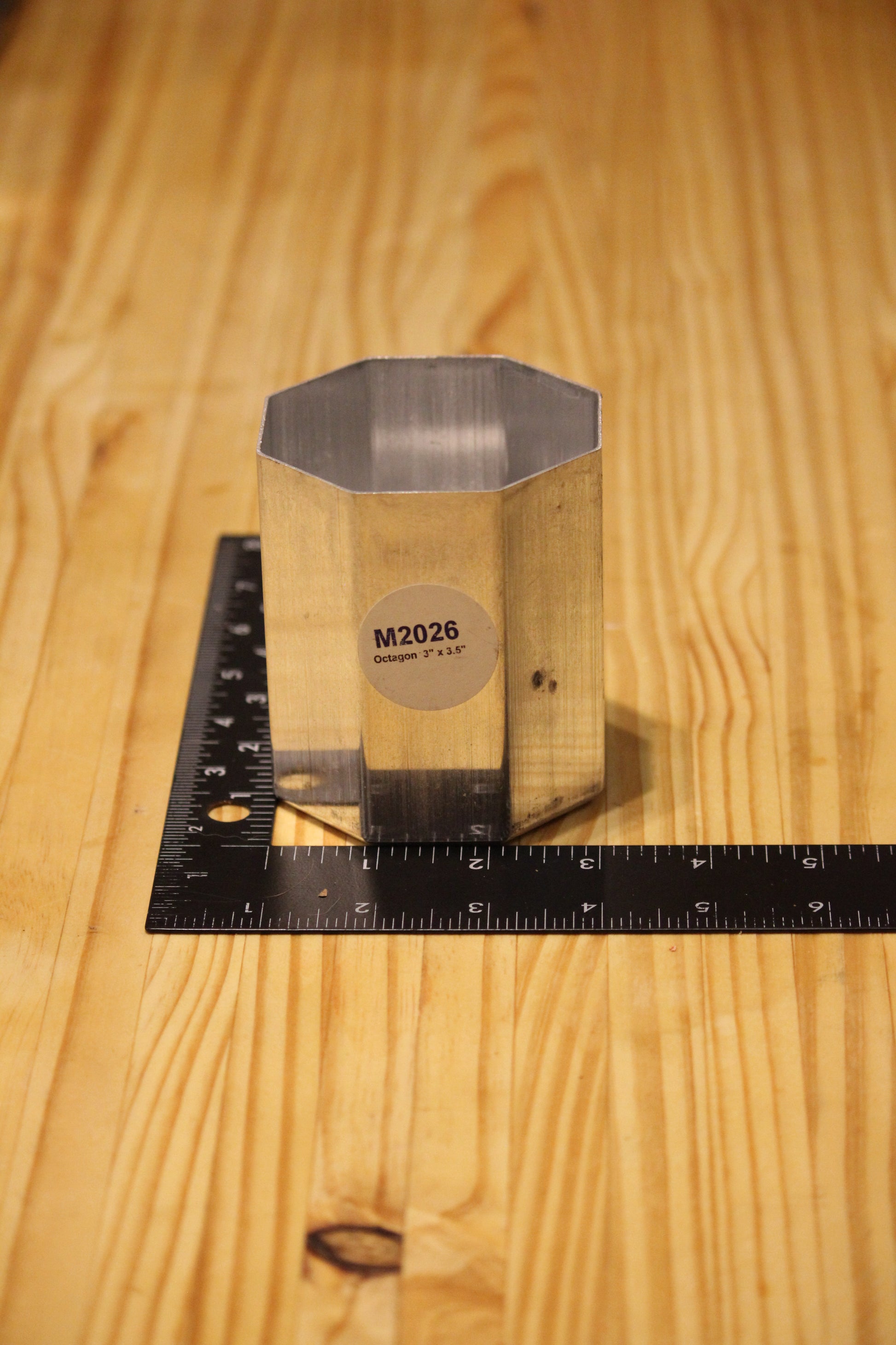 A small metallic octagonal block with a circular label reading 'M2026' stands on a black ruler on a light wood tabletop.