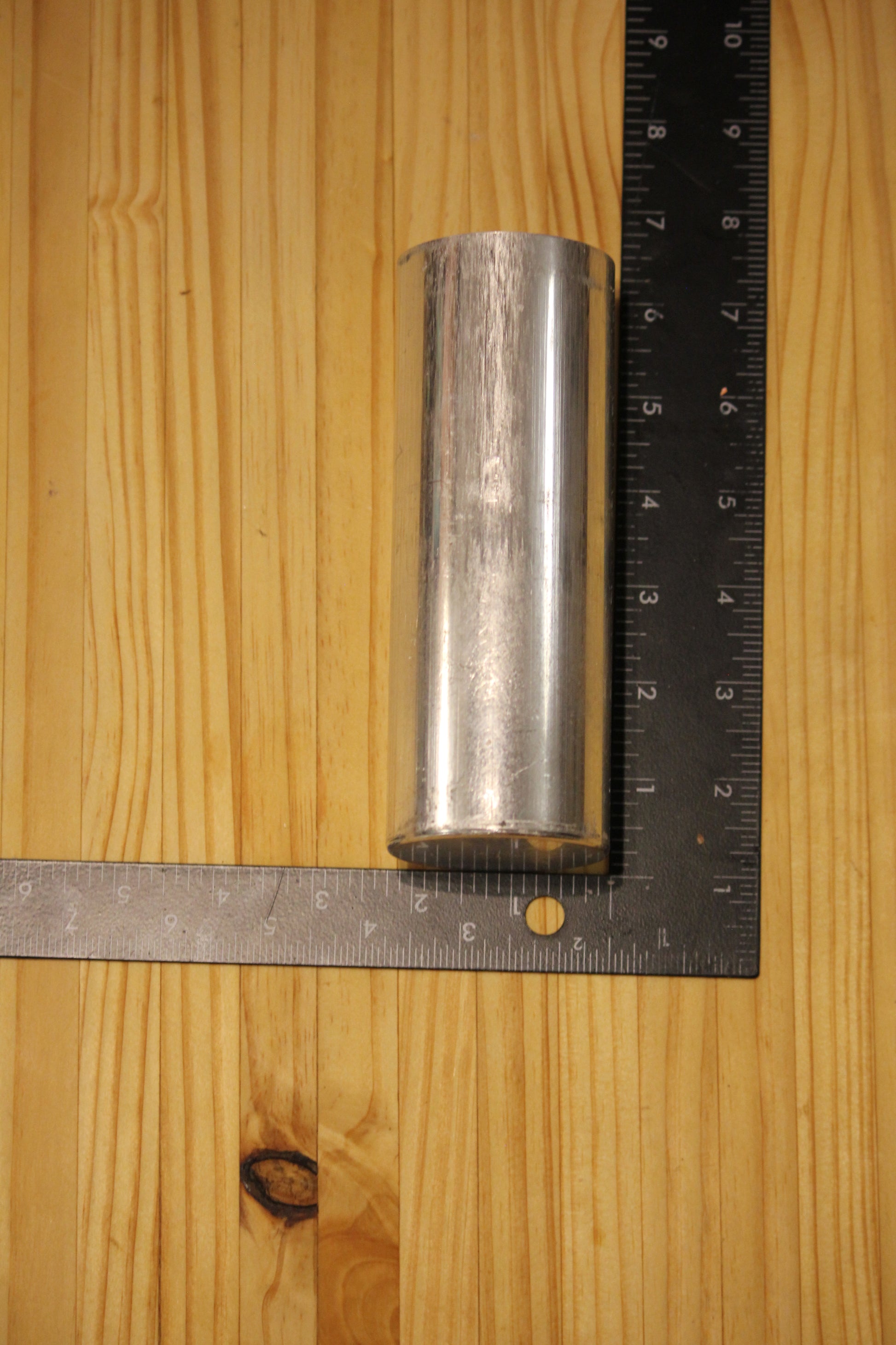 A small cylindrical metal pillar mold, shown on a light wood grain surface with a black ruler for scale.