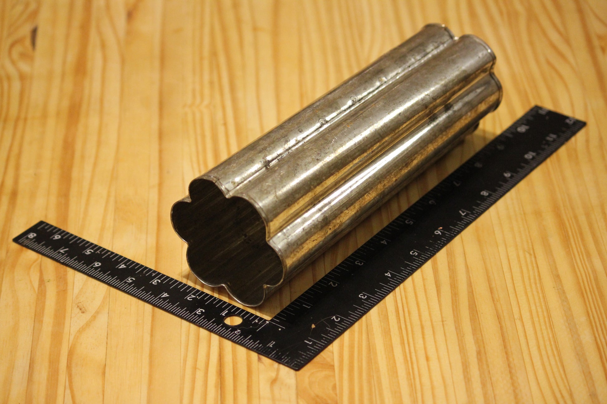 A metal flower shaped pillar mold, shown on a light wood grain background with a black ruler for scale.