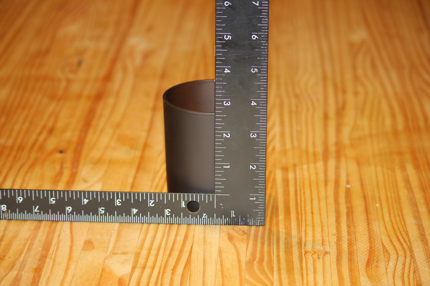 A dark brown candle vessel on a light wood grain surface, with metal rulers placed along the left and right sides to show its height in centimeters and inches.
