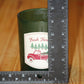A dark green glass jar candle with a 'Fresh Farm' label featuring a red truck and pine trees, shown next to a black metal ruler on a wooden surface to indicate size.