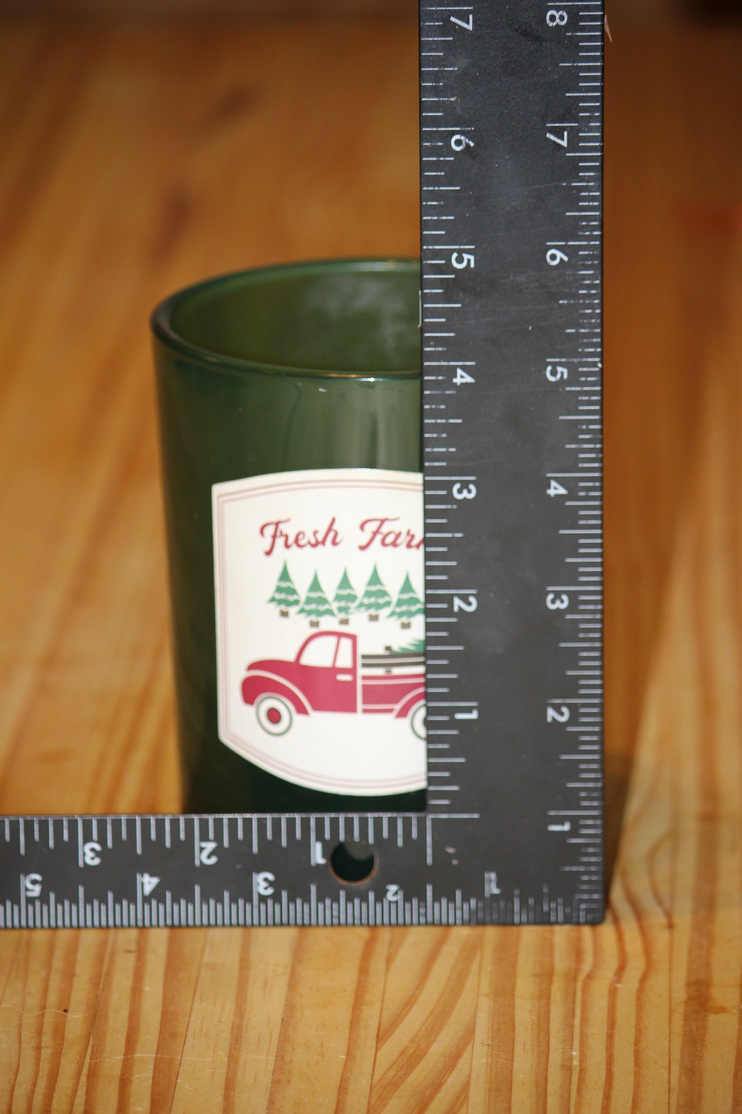 A dark green glass jar candle with a 'Fresh Farm' label featuring a red truck and pine trees, shown next to a black metal ruler on a wooden surface to indicate size.