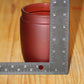 A small matte red candle holder shown on a wooden surface with a metal ruler standing beside it for scale.