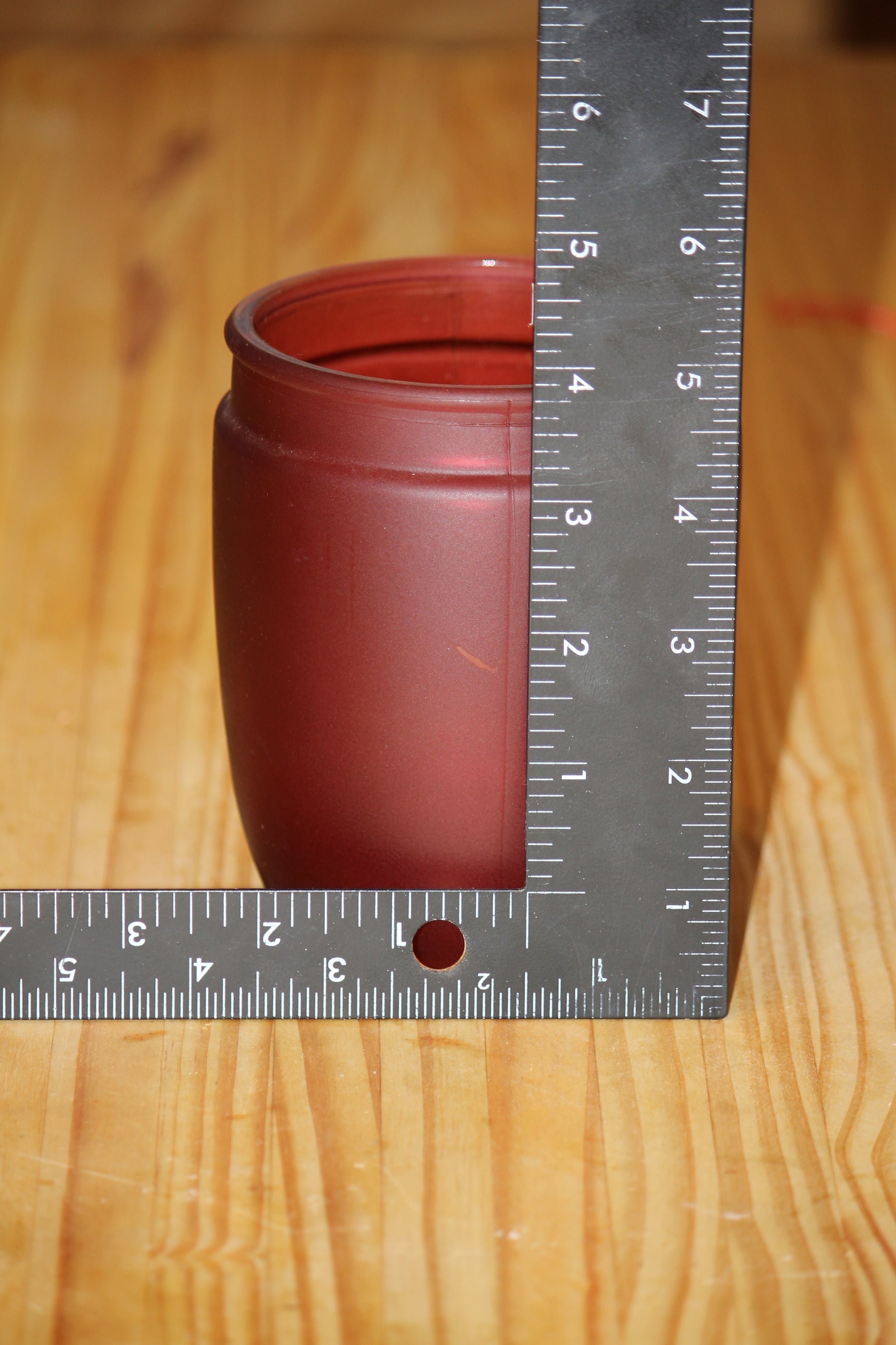 A small matte red candle holder shown on a wooden surface with a metal ruler standing beside it for scale.