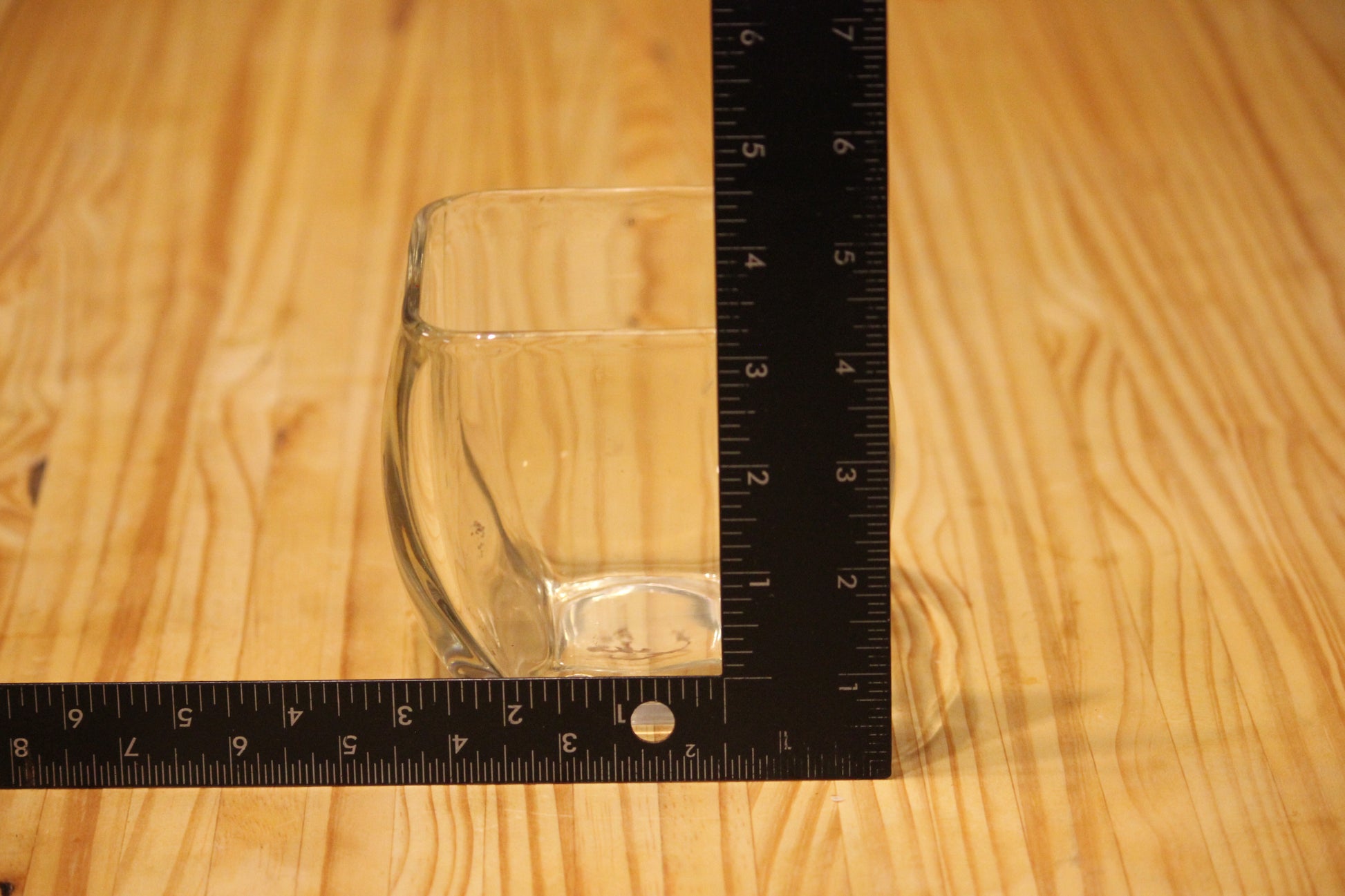 A clear glass candle vessel on a light wood surface with a black L-shaped ruler measuring its height and width.