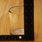 A clear glass candle vessel on a wooden surface with a black measuring ruler standing vertically and a second ruler lying horizontally to show its dimensions.