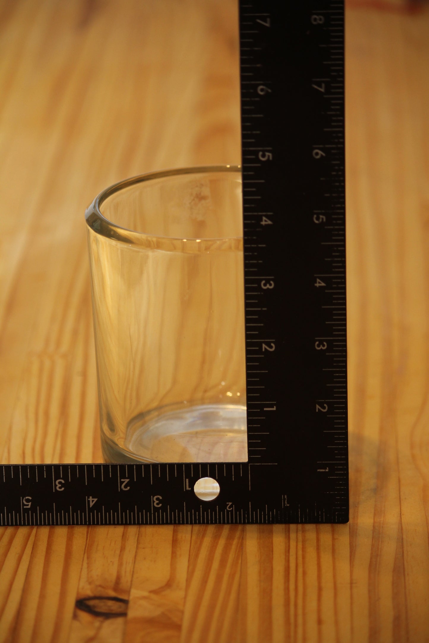 A clear glass candle vessel on a wooden surface with a black measuring ruler standing vertically and a second ruler lying horizontally to show its dimensions.
