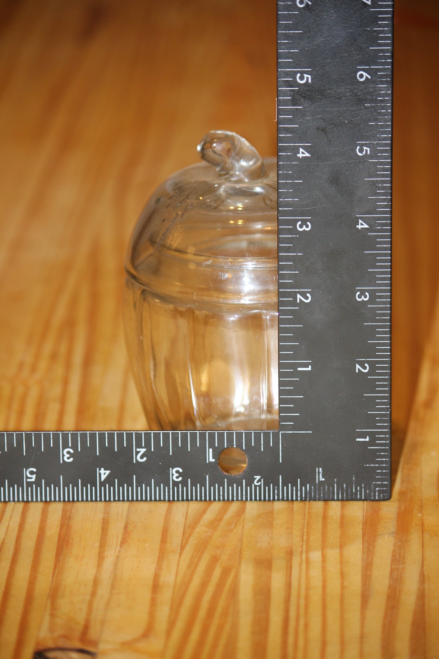 A small clear glass lidded apple jar with a knob on top, shown on a wooden surface with a metal ruler standing vertically and horizontally to indicate its size.