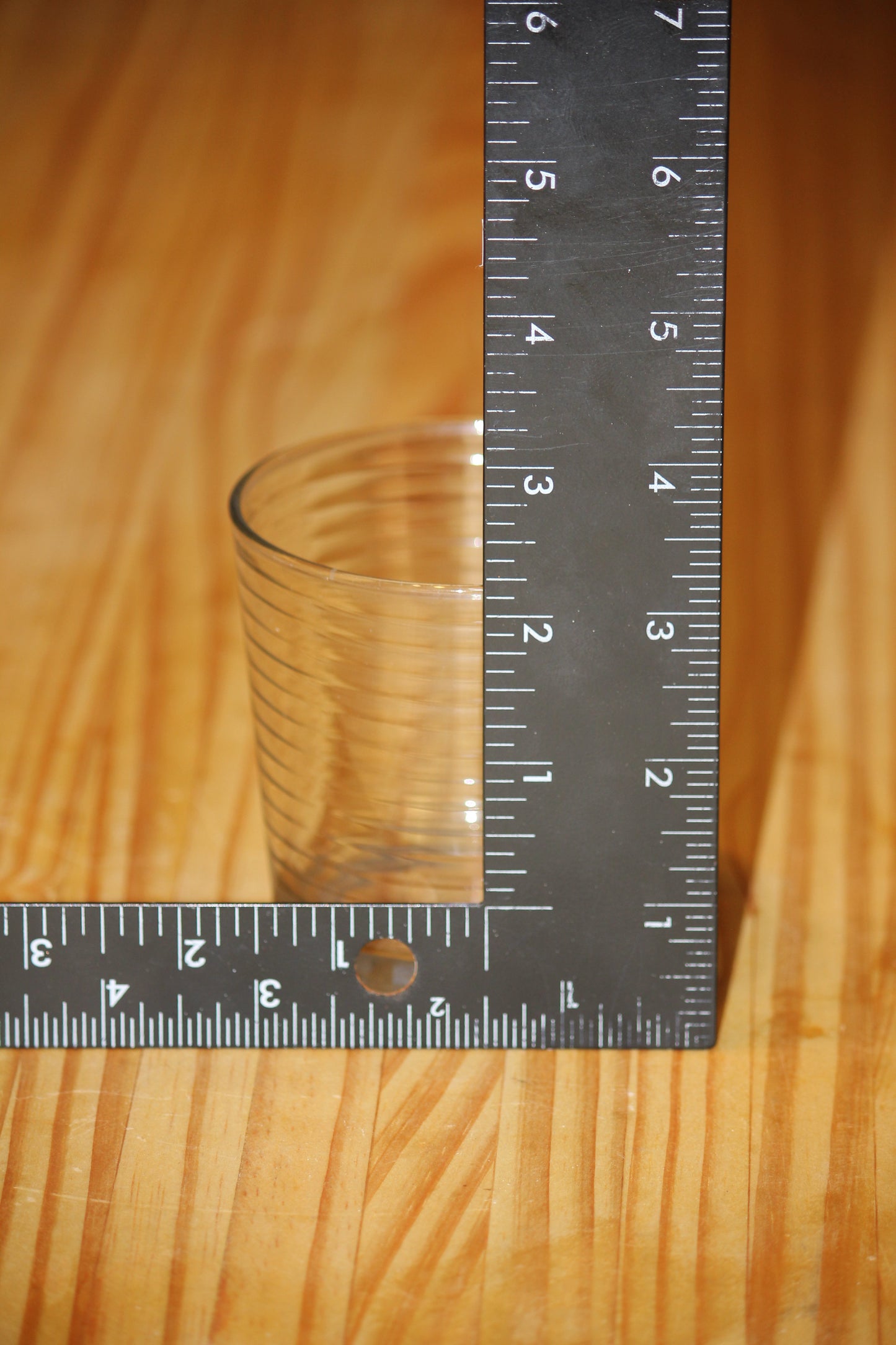 A clear ribbed glass candle vessel, standing on a wooden surface next to a black metal L-square ruler, showing its dimensions against a light wood grain background.
