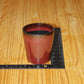 A small candle vessel in a reddish-brown glaze with a dark rim, shown on a wooden tabletop next to a black ruler for scale.