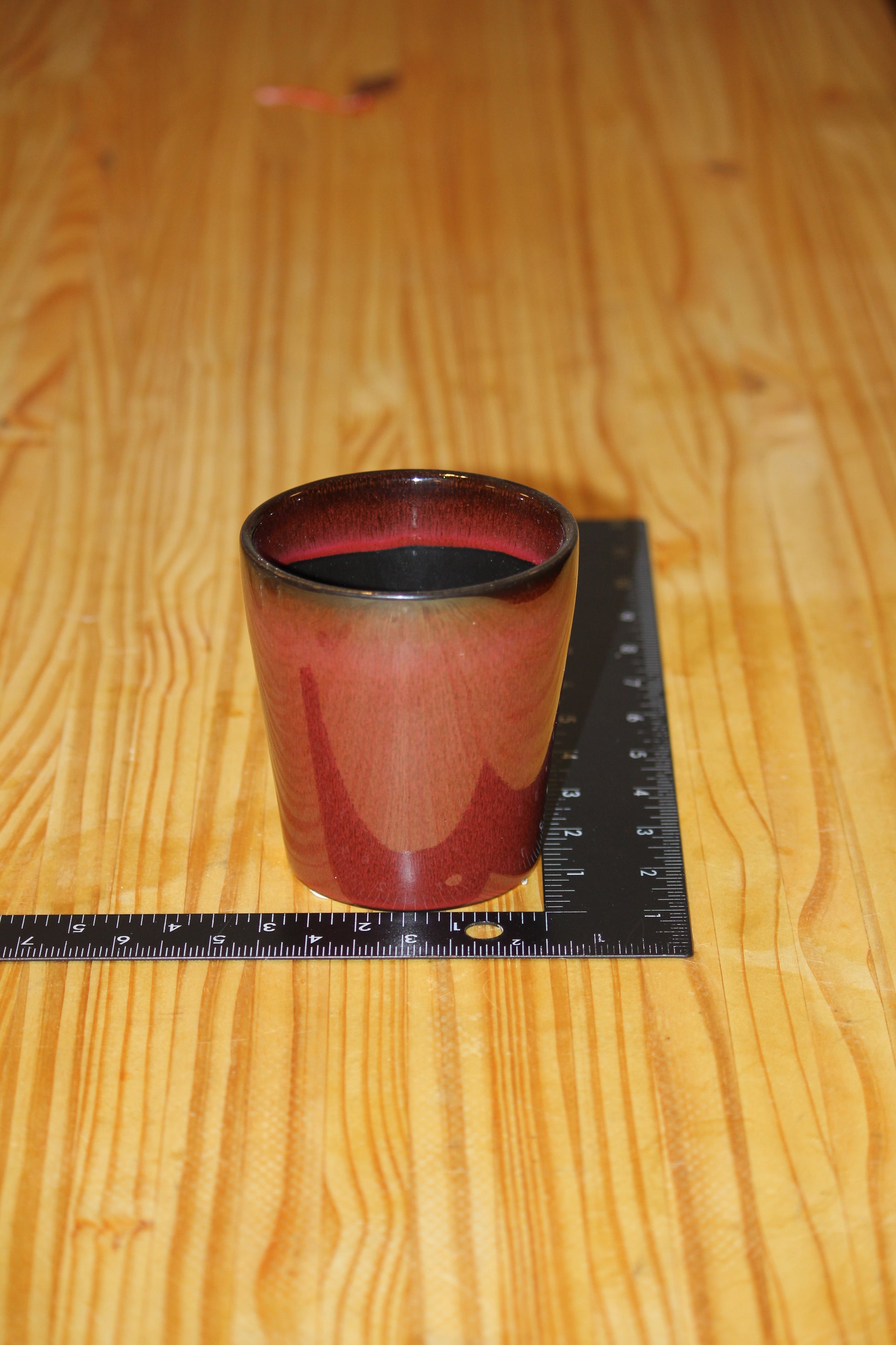 A small candle vessel in a reddish-brown glaze with a dark rim, shown on a wooden tabletop next to a black ruler for scale.