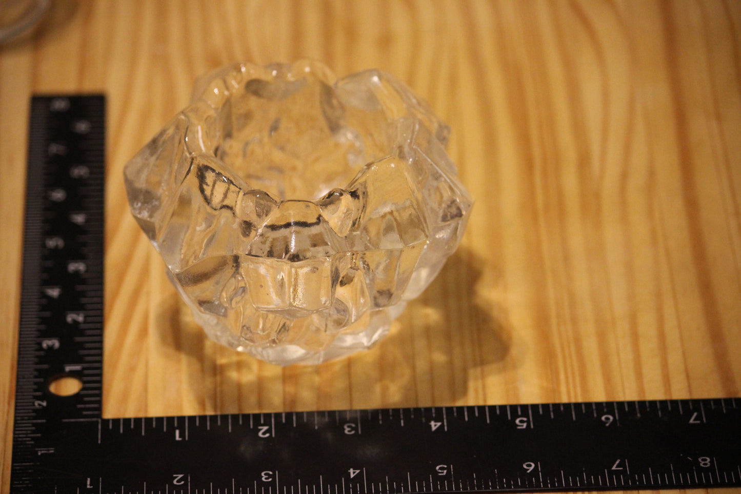 A small clear faceted glass candle holder placed on a wooden surface with a black ruler visible along the left and bottom edges for scale.